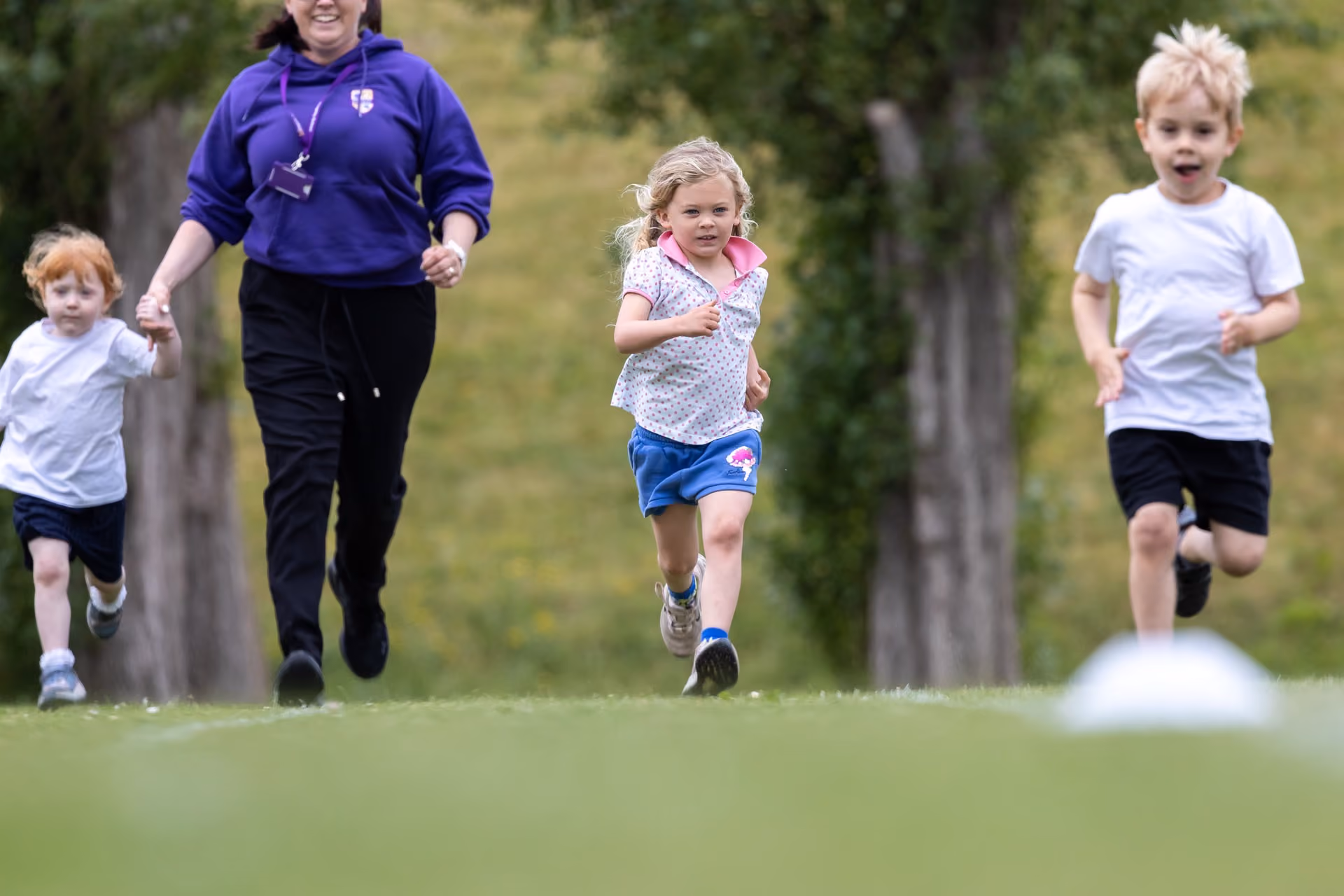 Three young children and an adult running on a grassy field, with trees in the background, during an outdoor activity or sports day.