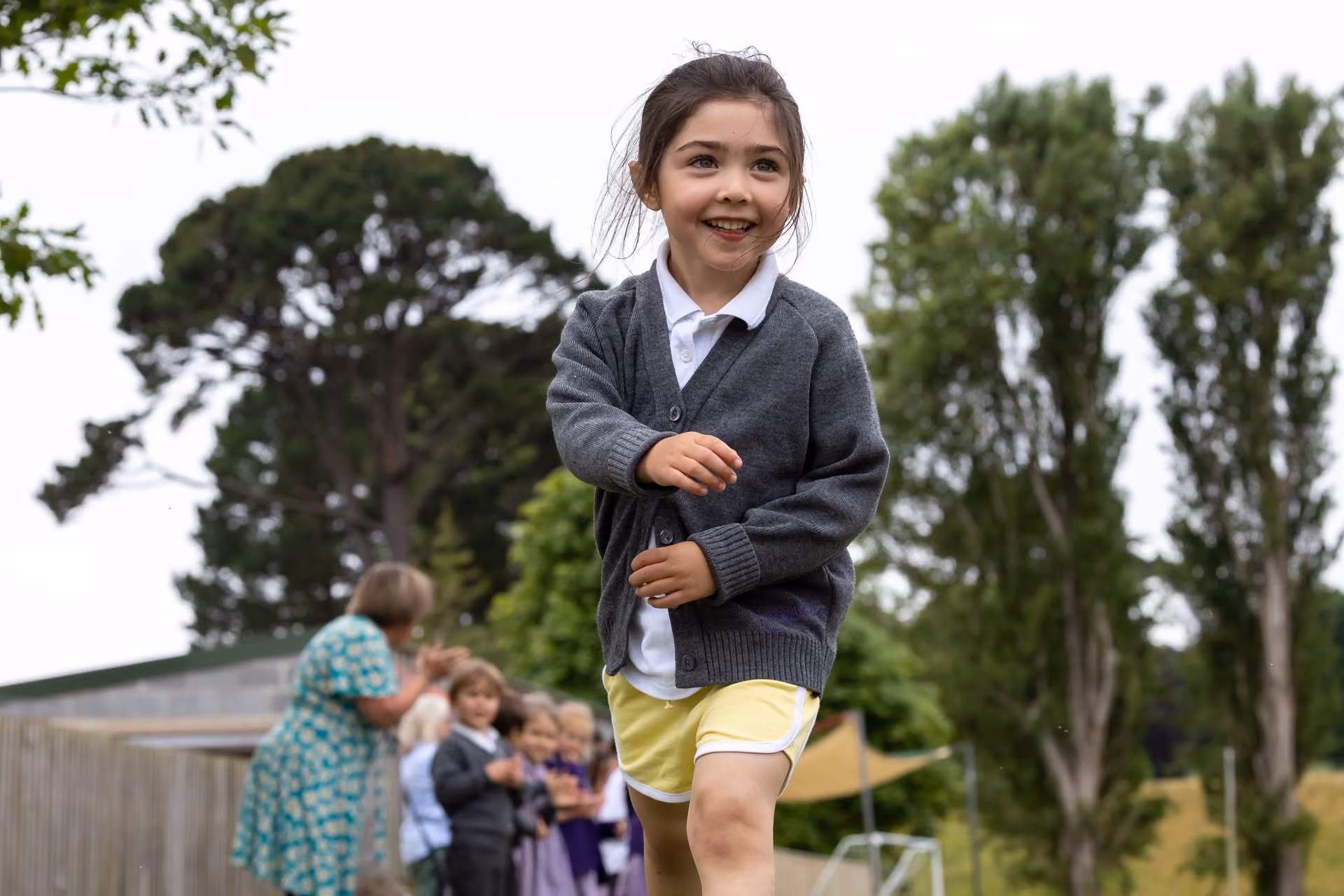 Smiling young girl in a grey cardigan and yellow shorts running outdoors on a grassy field during a school sports day, with adults and children blurred in the background near trees.