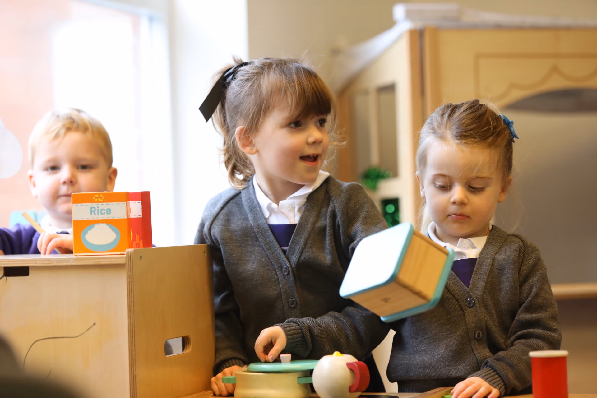 Three young children in school uniforms and grey cardigans playing with toy kitchen items in a classroom, including a block, a toy pot, and a cereal box labelled Rice.
