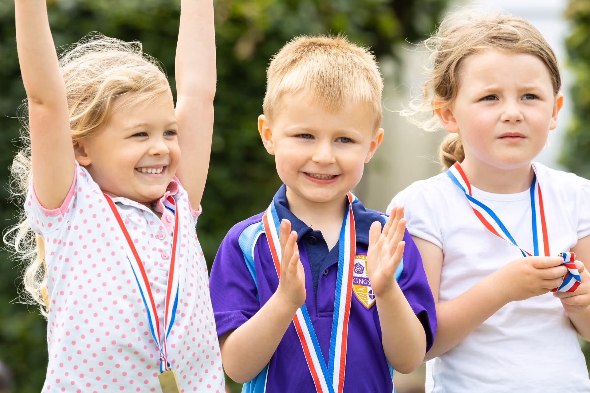 Three young children wearing medals around their necks, one girl raising her arms excitedly, a boy clapping, and another girl holding her medal with a thoughtful expression, outdoors with greenery in the background.