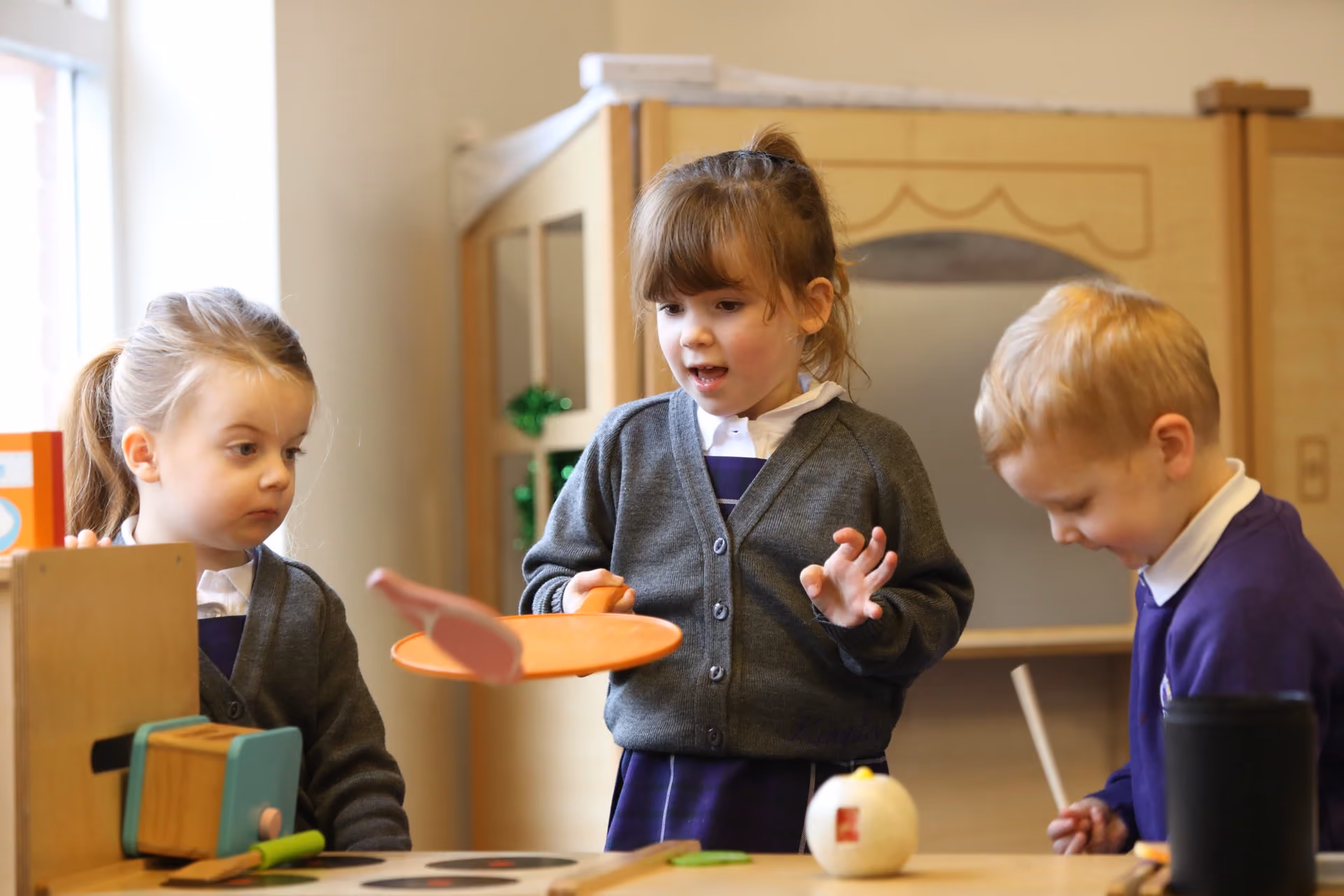 Three young children in school uniforms playing with toys at a table indoors, with one girl holding an orange toy plate and another boy focused on playing.
