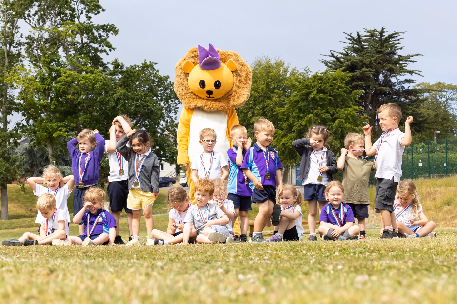 Group of young children sitting and standing outdoors on grass wearing sports day medals, with a person dressed in a large lion mascot costume with a purple party hat standing behind them, trees and a clear sky in the background.