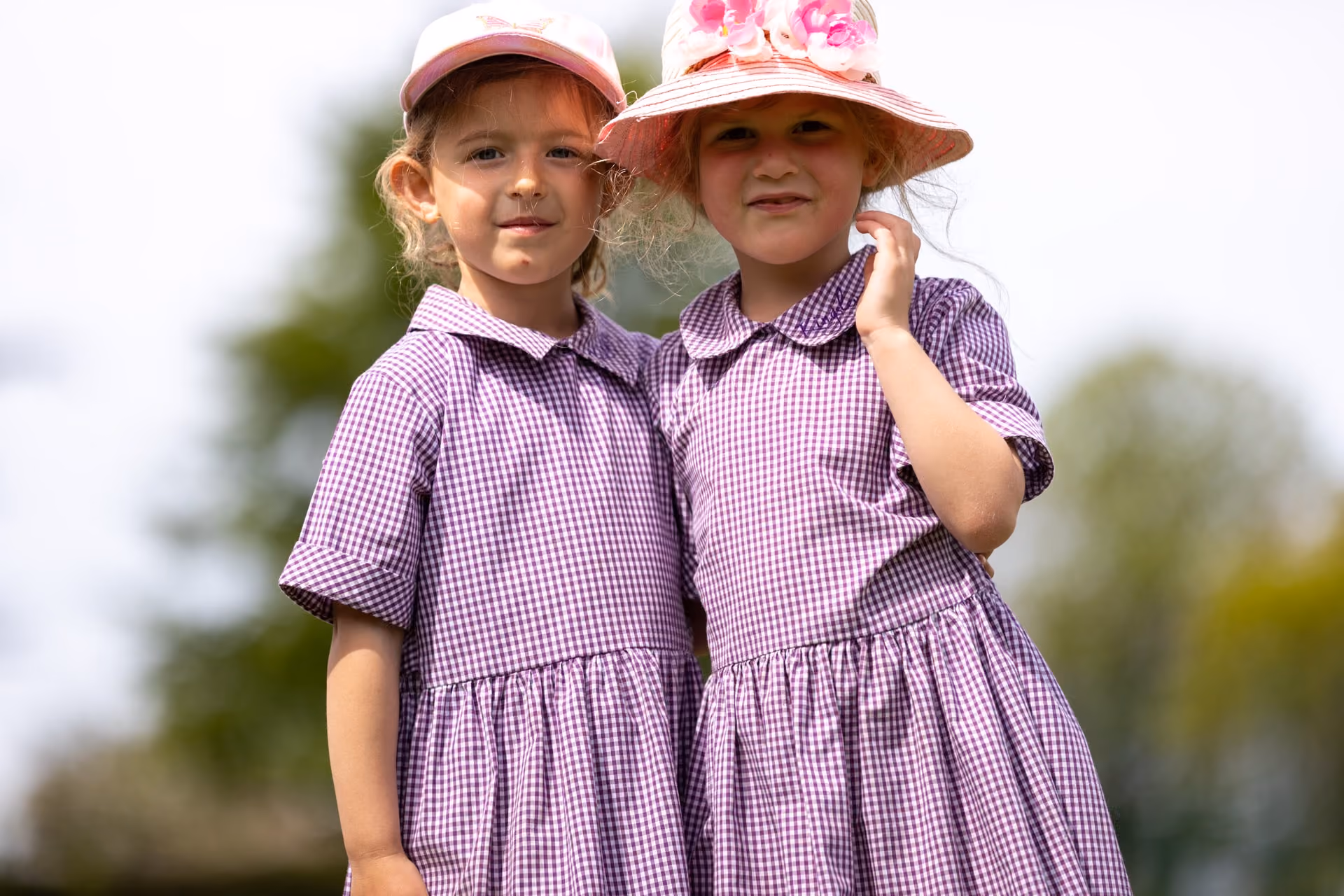 Two young girls wearing purple checkered dresses and sun hats stand close together outdoors with a blurred tree background.