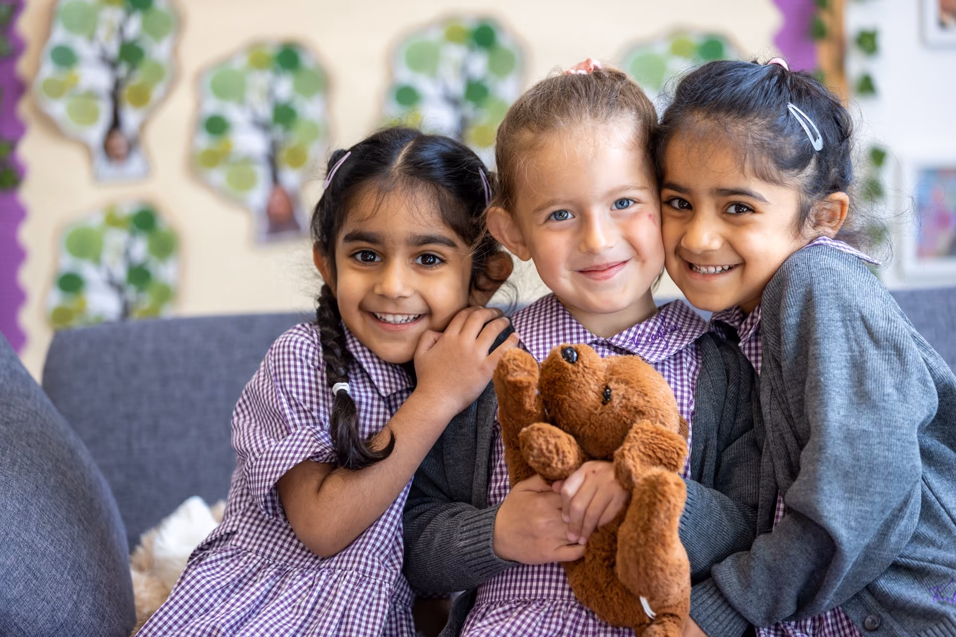 Three young girls in checked school uniforms and grey cardigans sitting closely together and smiling, one holding a brown teddy bear, in a classroom with colorful tree decorations on the wall behind them.