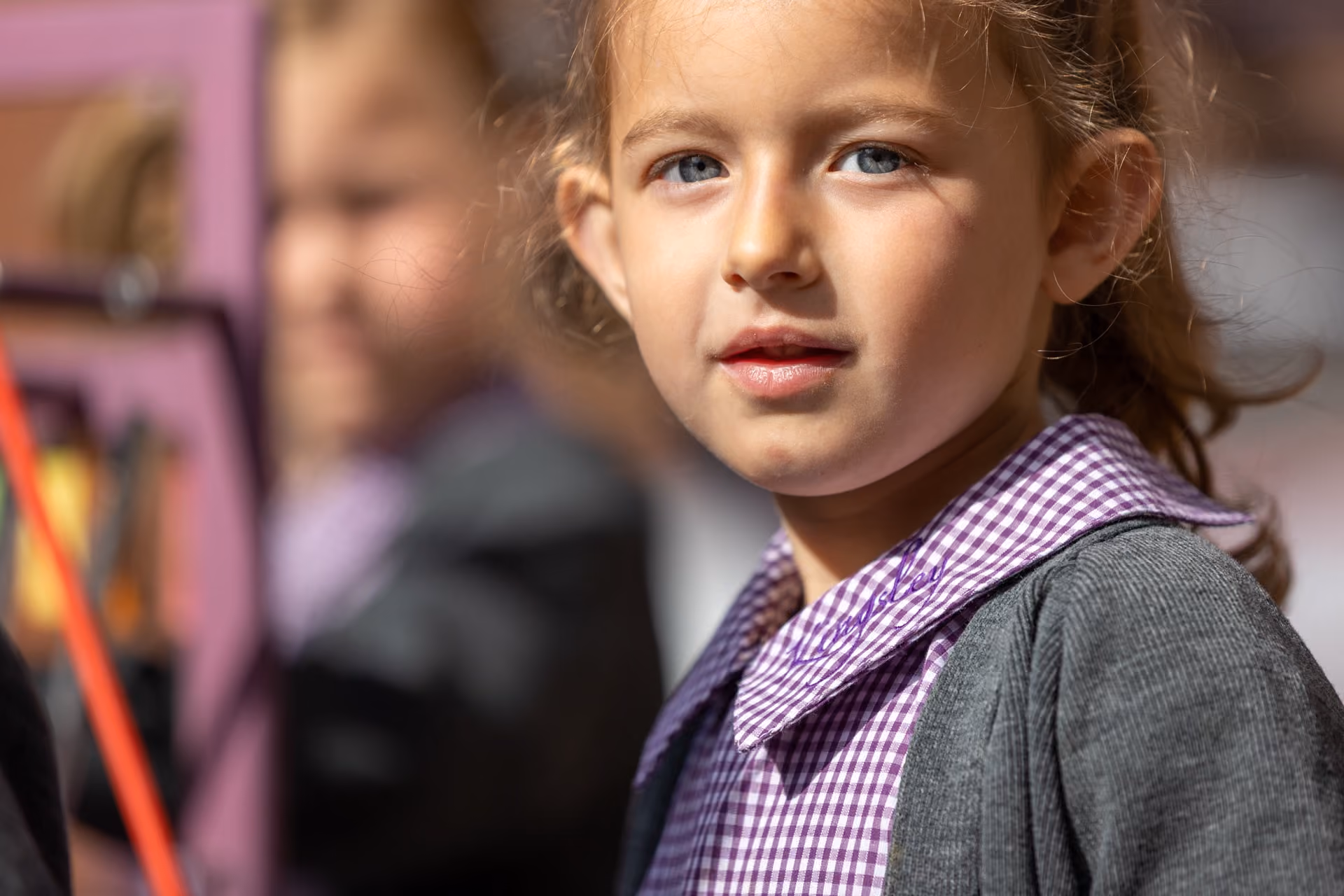 Close-up of a young girl with light brown hair and blue eyes wearing a purple checkered school uniform shirt and grey cardigan, looking at the camera with a neutral expression.