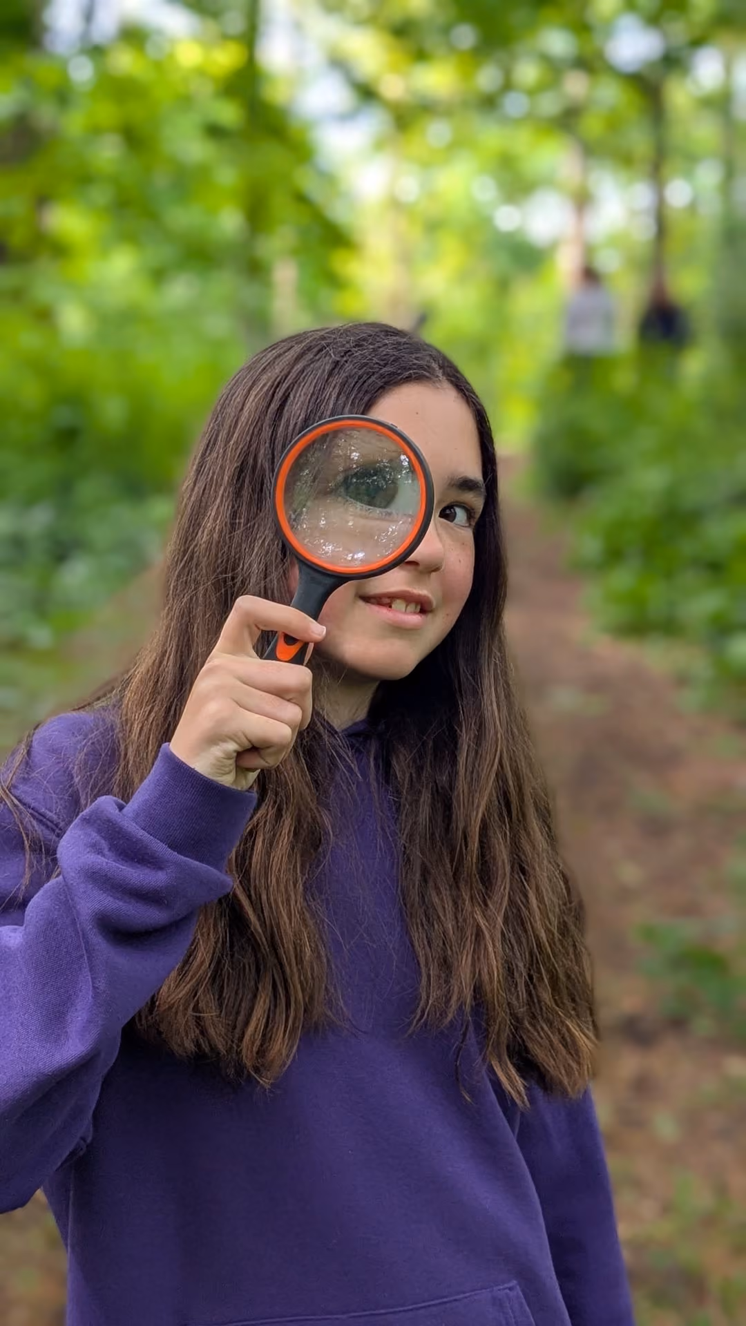 Young girl with long brown hair wearing a purple sweatshirt holds a magnifying glass up to her eye while standing on a leafy forest path.