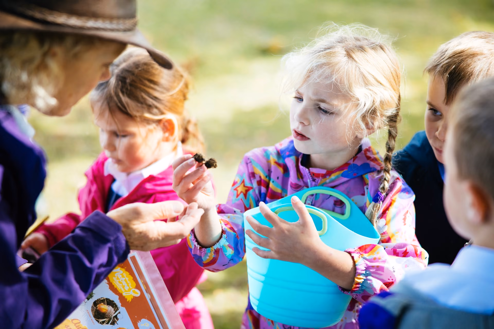 A young girl in a colourful jacket holds a blue basket and shows a small object to an adult wearing a hat, while other children watch outdoors on a sunny day.