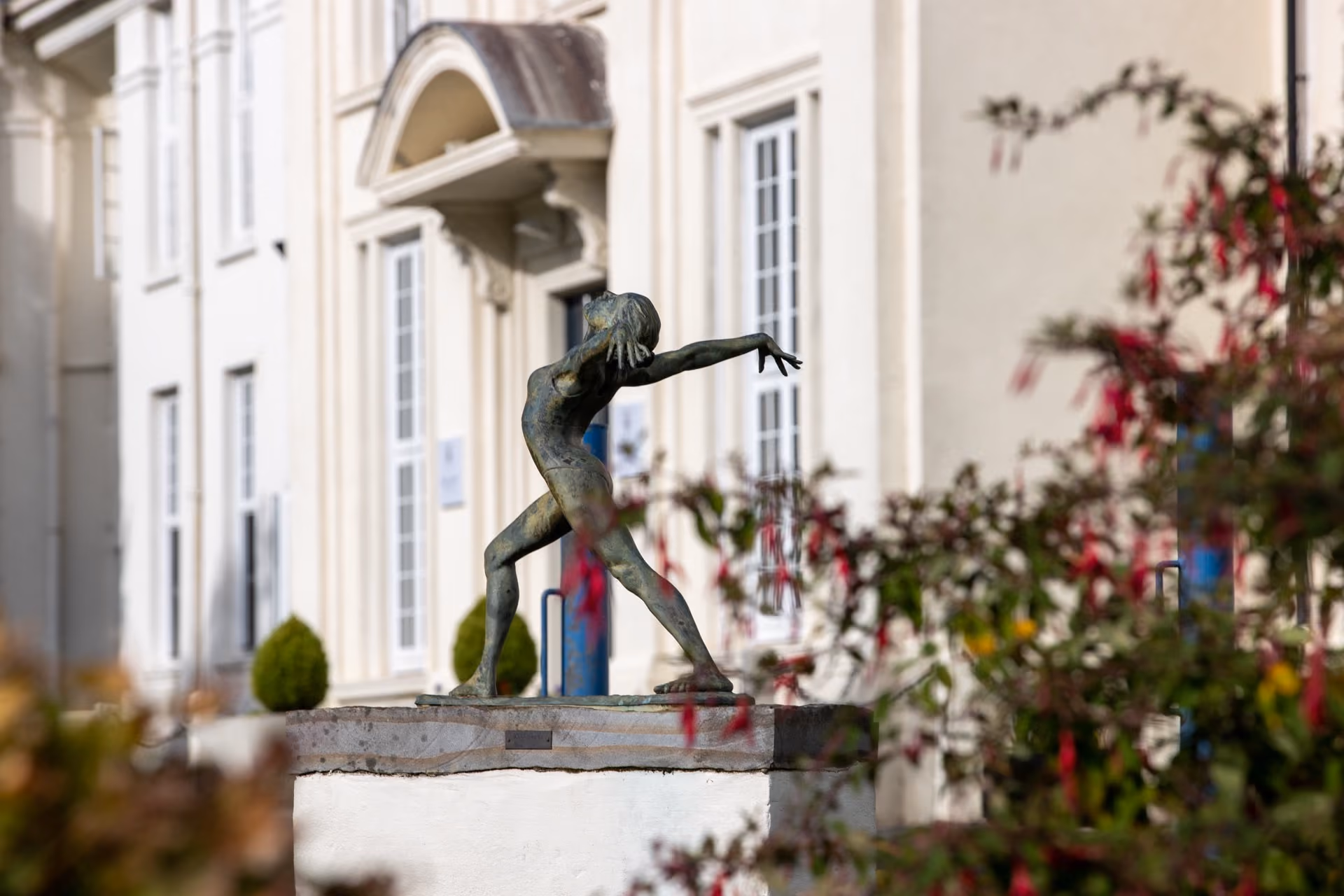 Bronze statue of a dancer with outstretched arms and bent knees on a pedestal, framed by blurred pink flowers, with a cream building featuring arched doorway and multiple tall windows in the background.