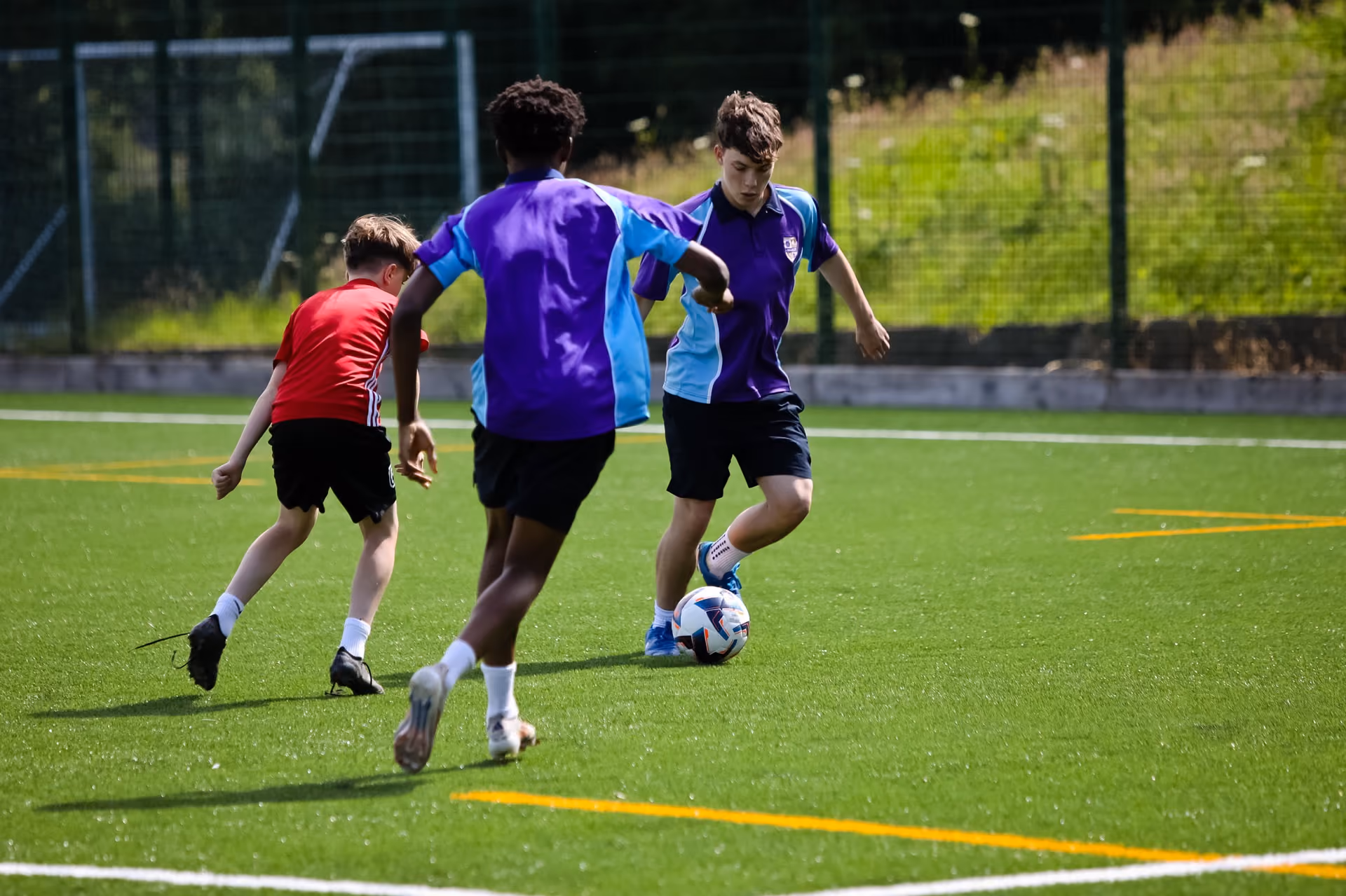 Three boys playing football on a green artificial pitch, with two boys in purple and blue kits and one boy in a red shirt chasing the ball.