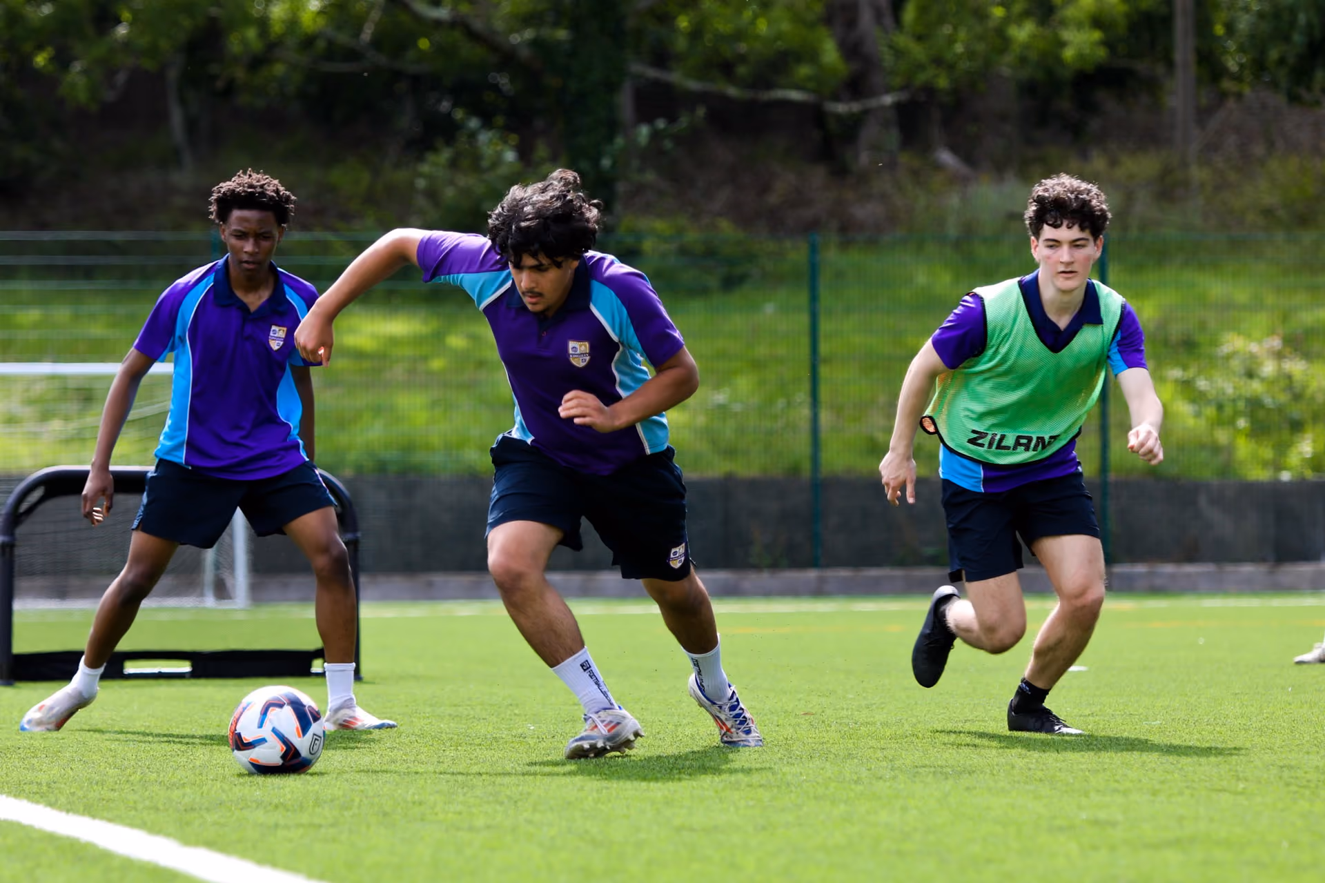 Three teenage boys playing football on a green artificial grass pitch, two boys wearing purple and blue sports shirts, one wearing a green bib, with one boy running towards the ball.
