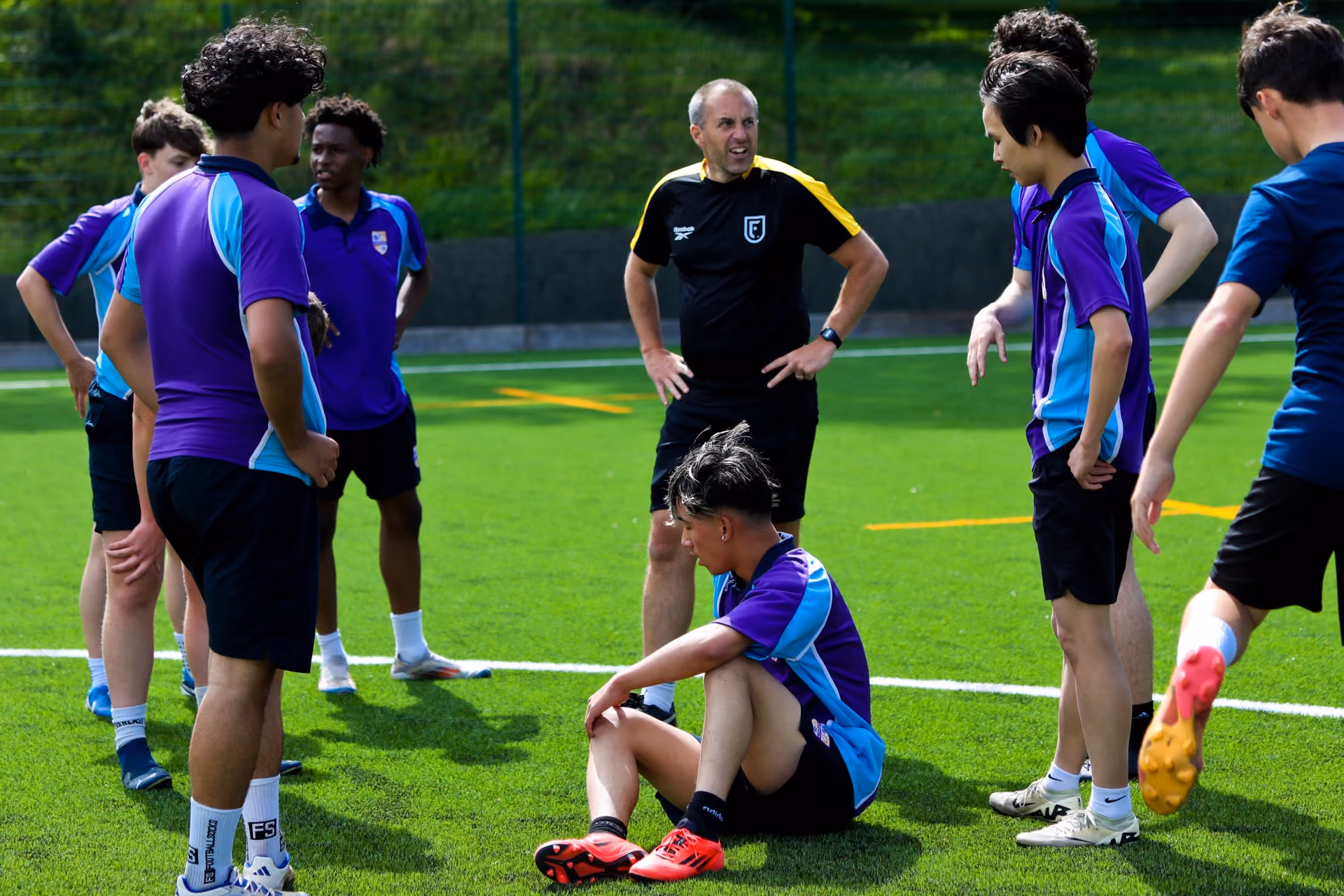 A football coach in black sportswear speaking to a group of young male players wearing purple and blue training kits on a green artificial turf field, with one player sitting on the ground and others standing around him.
