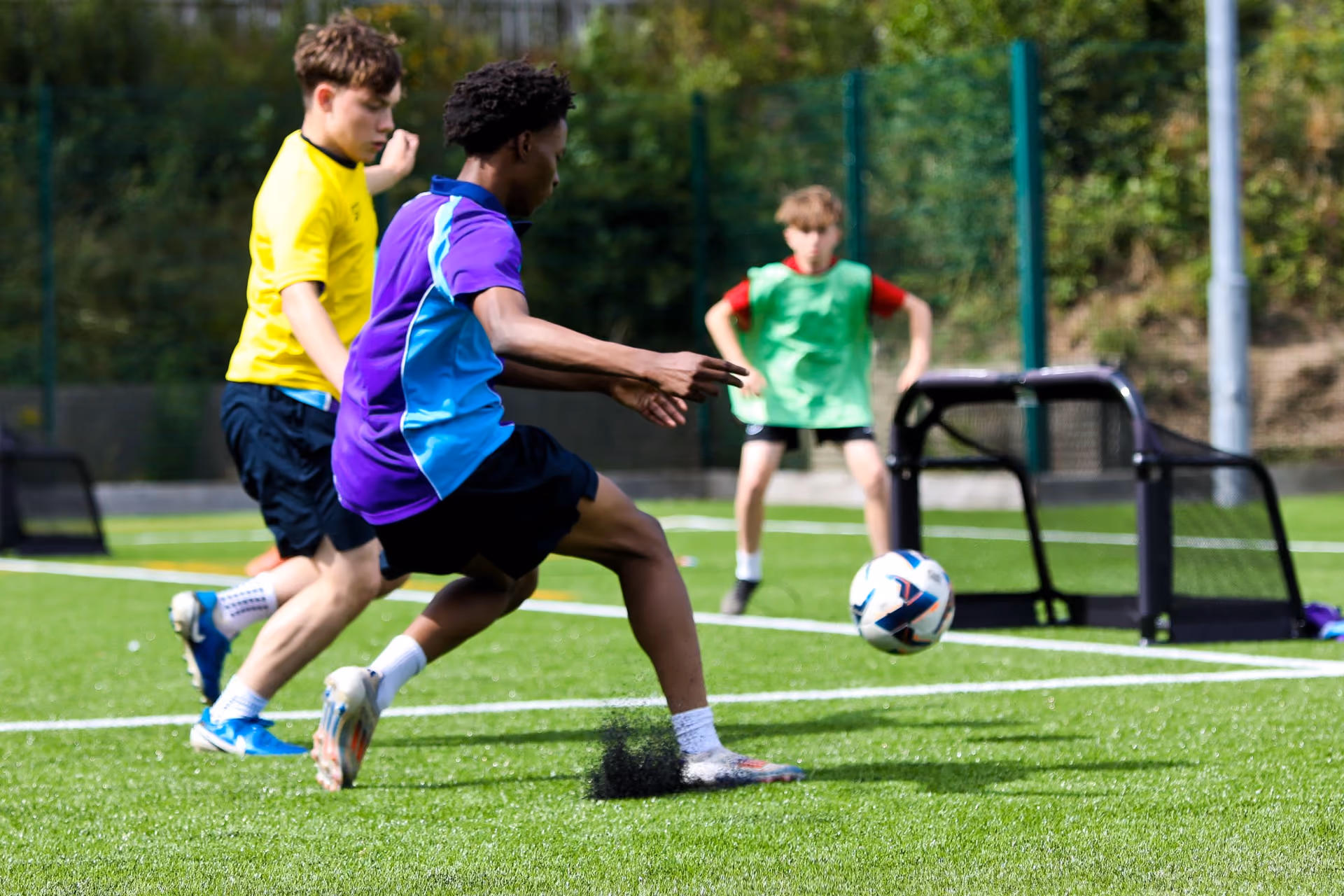 Three boys playing football on a green artificial turf pitch, one in a purple and blue shirt kicking the ball, another in a yellow shirt running behind, and a third boy in a green vest standing near a small goal in the background.
