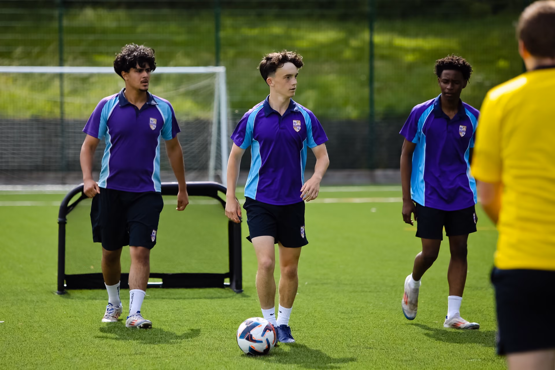 Three young male football players in purple and light blue jerseys walking on a green artificial turf field, one in the centre controlling a football, with a coach or referee in a yellow shirt partially visible in the foreground.