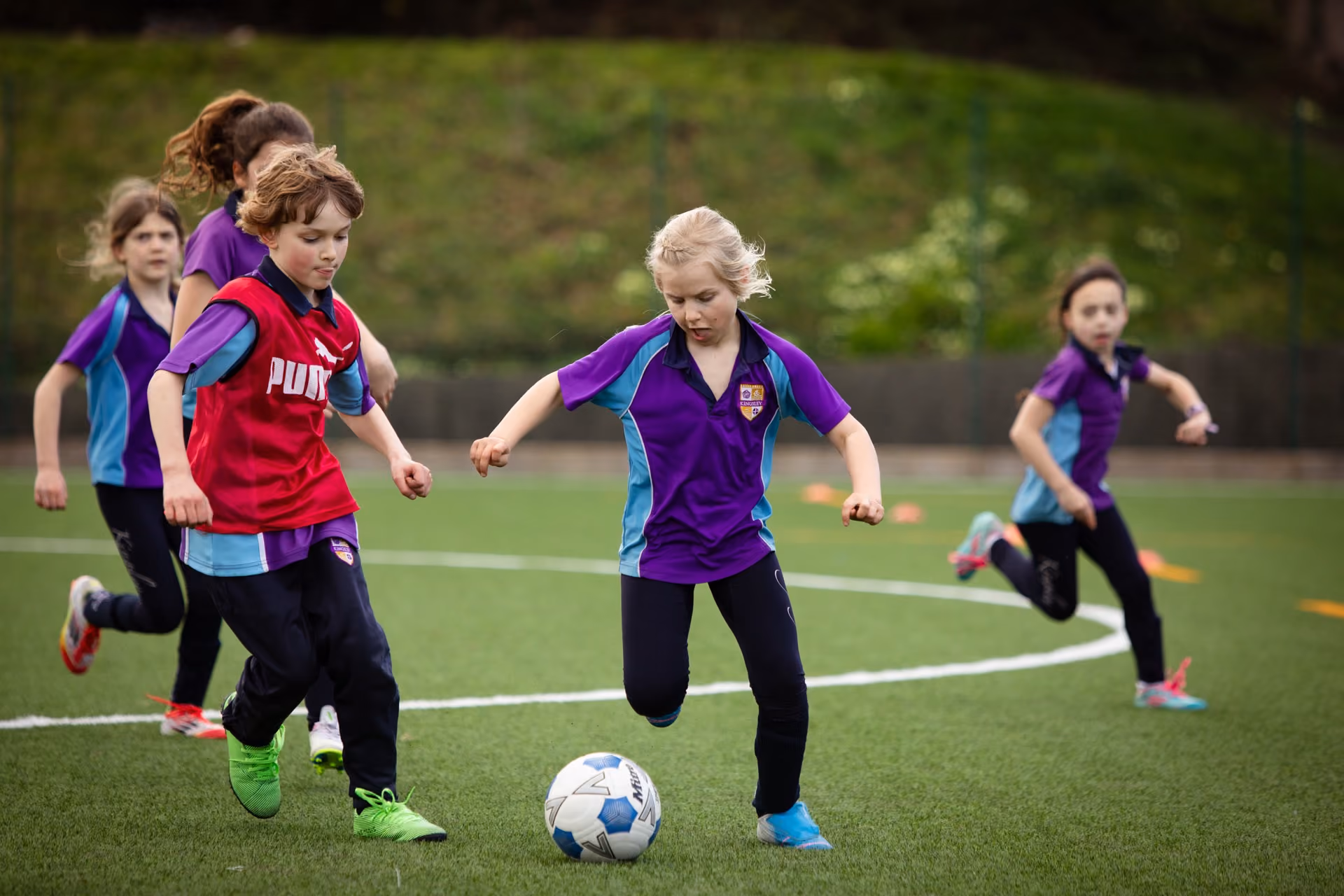 Young girls playing football on a grass field, with one girl in a red bib and green boots running towards the ball while another girl in a purple and blue kit runs alongside her, others in similar kits are seen in the background.