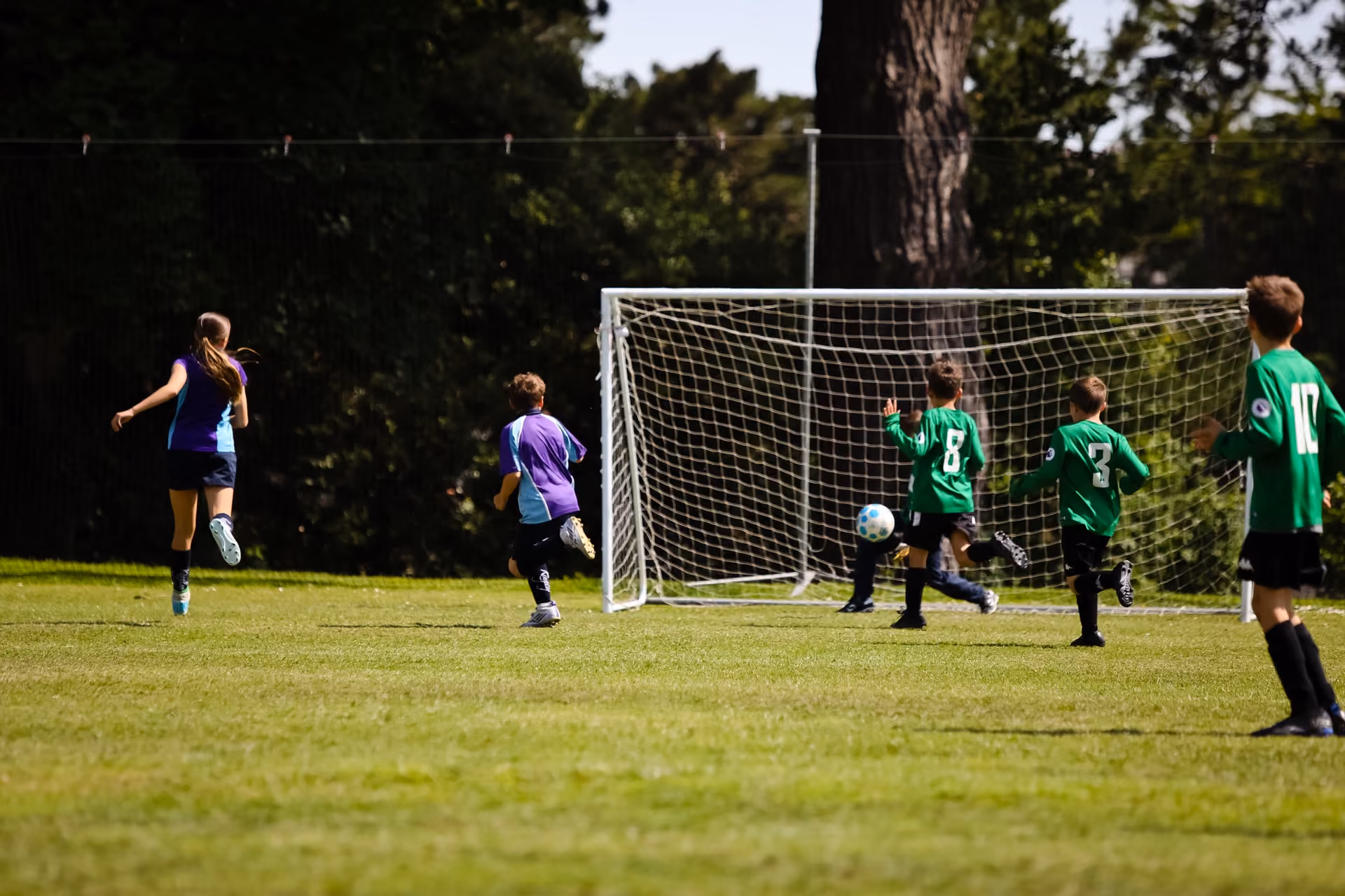 Children playing soccer on a grass field with one team wearing green jerseys and the other in purple, a ball near the goalpost.
