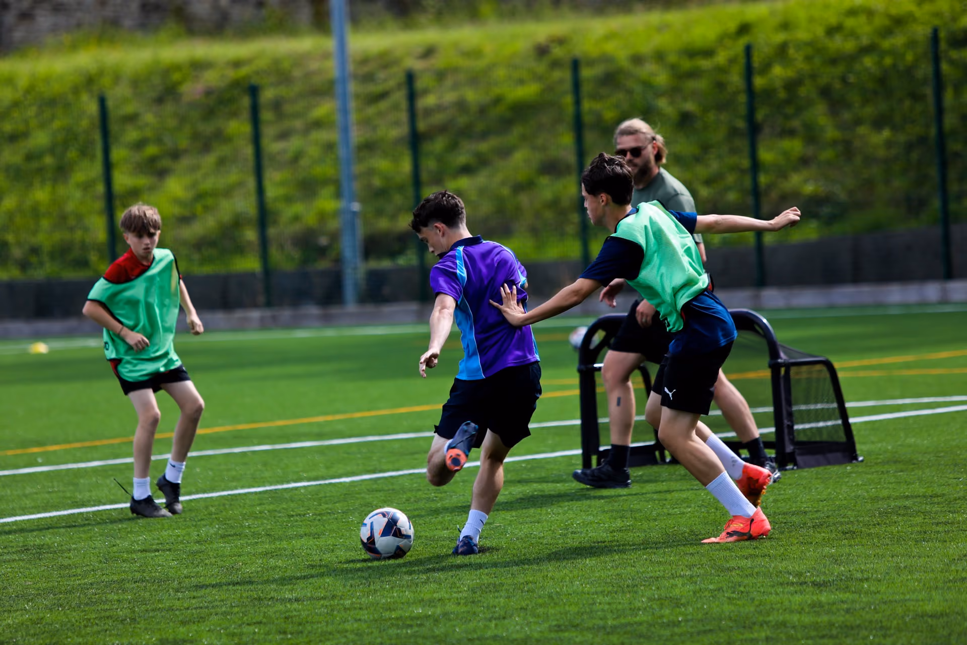 Three boys playing football on a grass pitch with one kicking the ball and another trying to block him, a coach or adult stands nearby watching.