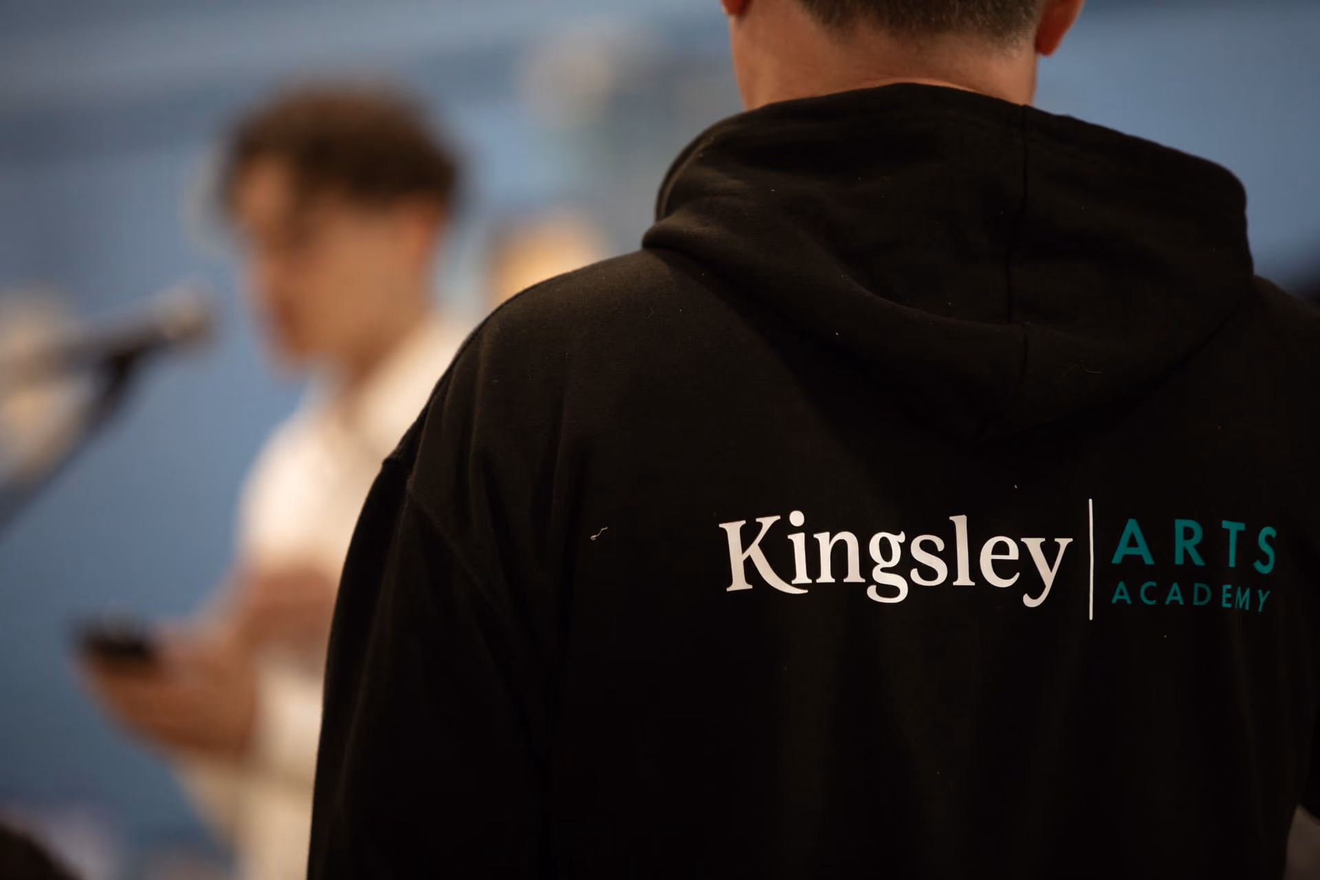 Person wearing a black hoodie with Kingsley Arts Academy logo on the back, blurred figure with microphone in the background.