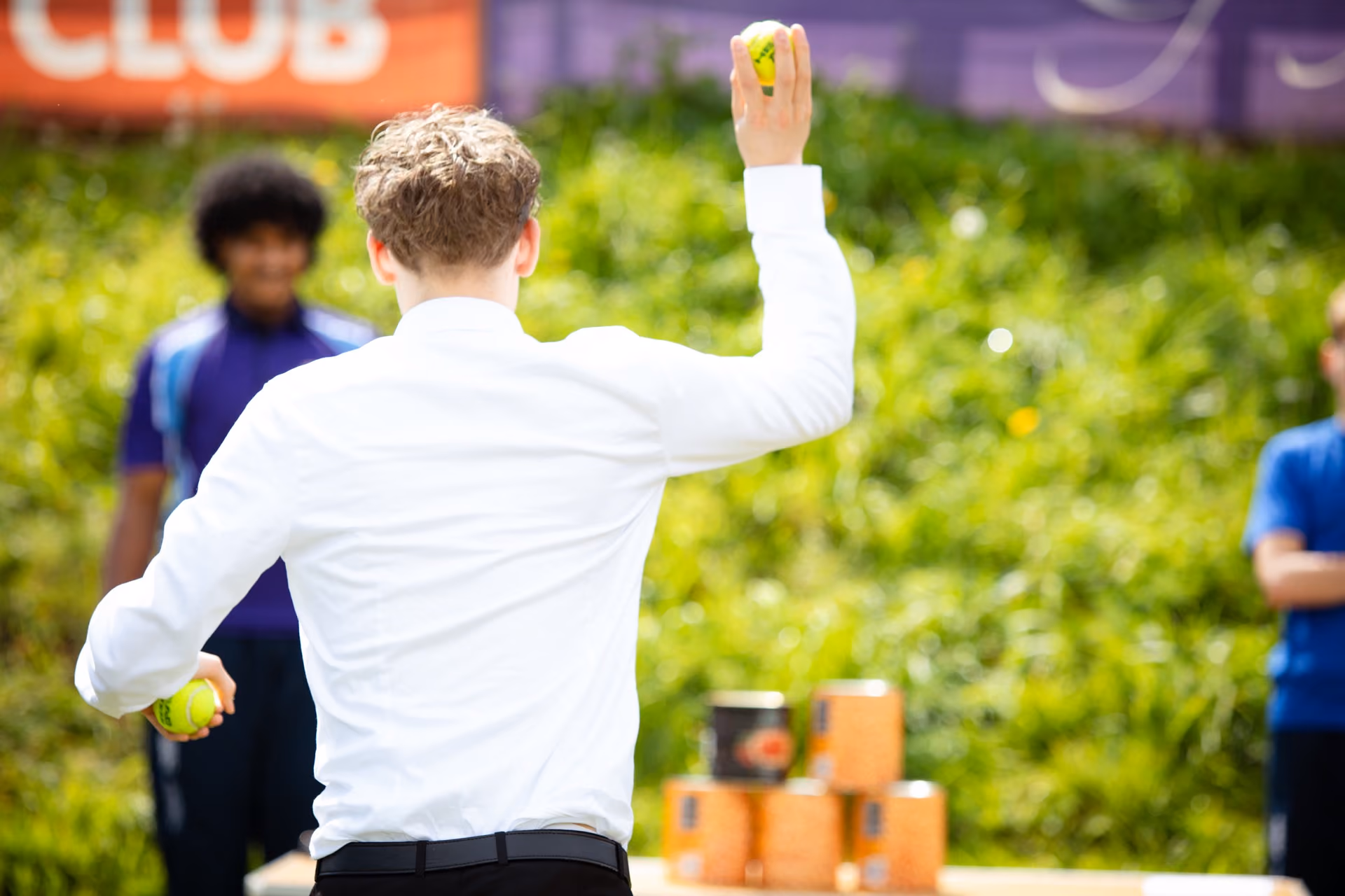 Person in a white shirt throwing a ball at a stack of orange cans in an outdoor game, with two onlookers in the background.