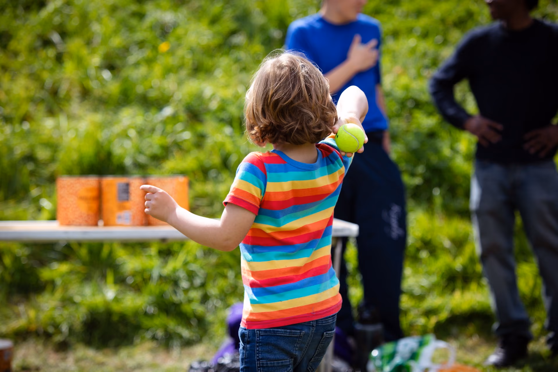 Child in a rainbow-striped shirt holding a tennis ball, preparing to throw it outdoors with two adults in the background near a table with orange boxes.