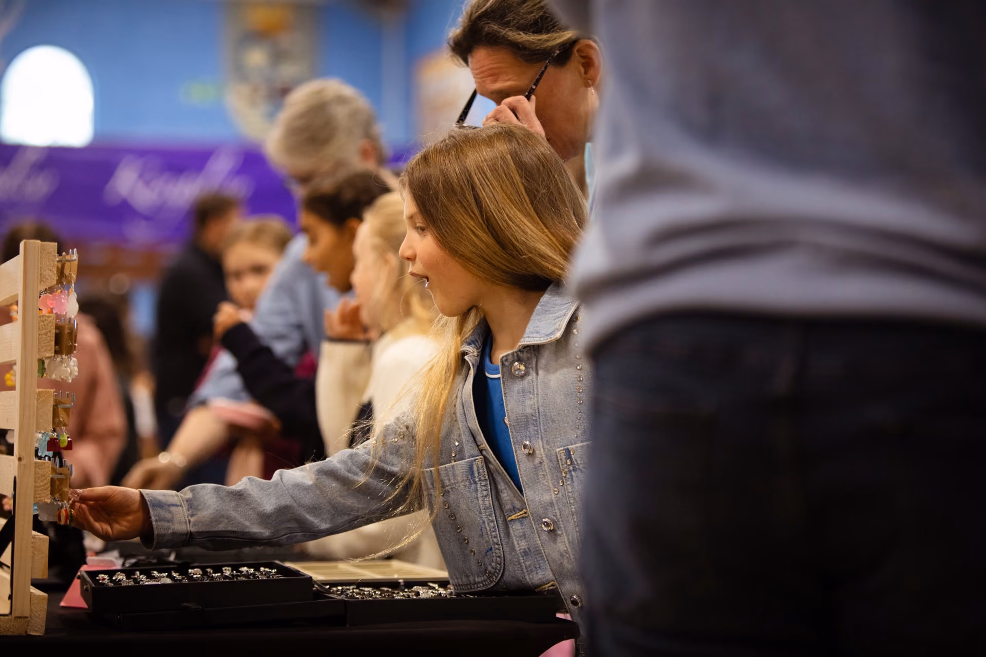 A young girl with long blonde hair wearing a denim jacket reaches out to select a piece of jewelry from a display at a busy indoor market or event with other people browsing in the background.