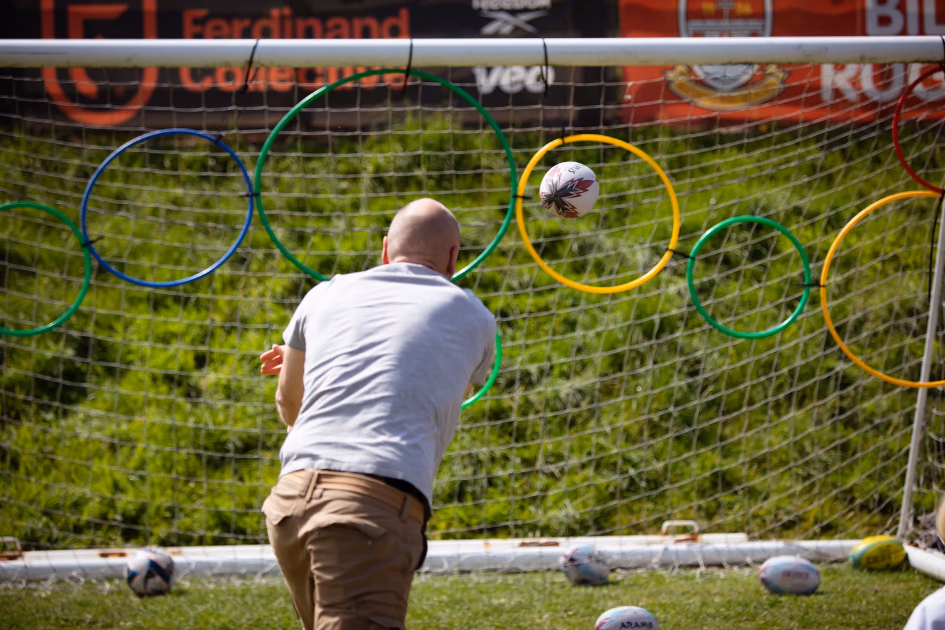 Man in white shirt and beige trousers throwing a ball through one of several colorful hoops attached to a football goal net outdoors.