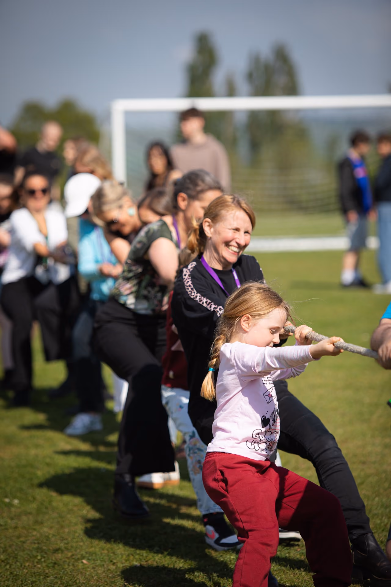 A group of children and adults playing tug-of-war outdoors on grass, with a young girl in a pink top and red trousers in the foreground pulling the rope, smiling adults behind her, and a football goal visible in the background.