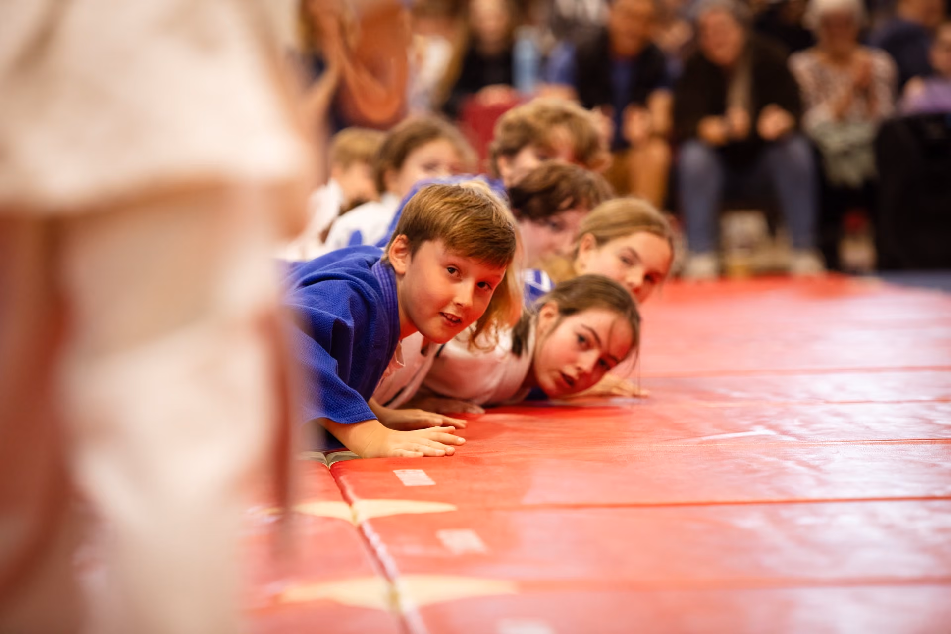 Children in judo uniforms lying on their stomachs on a red mat, looking intently toward the left side of the image with blurred spectators in the background.