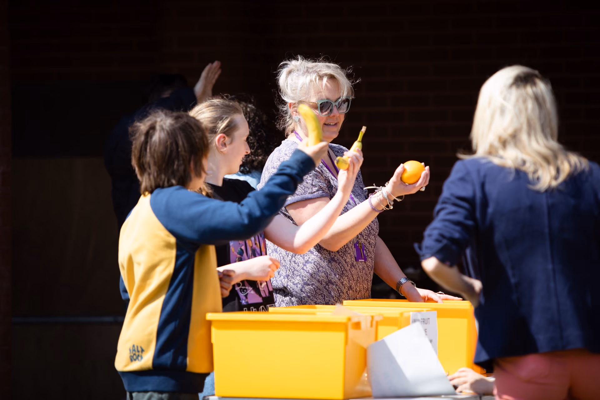 A group of people, including two children and two adults, stand around bright yellow plastic boxes, holding bananas and an orange. The woman with short white hair and sunglasses is smiling and appears to be engaging with the others.