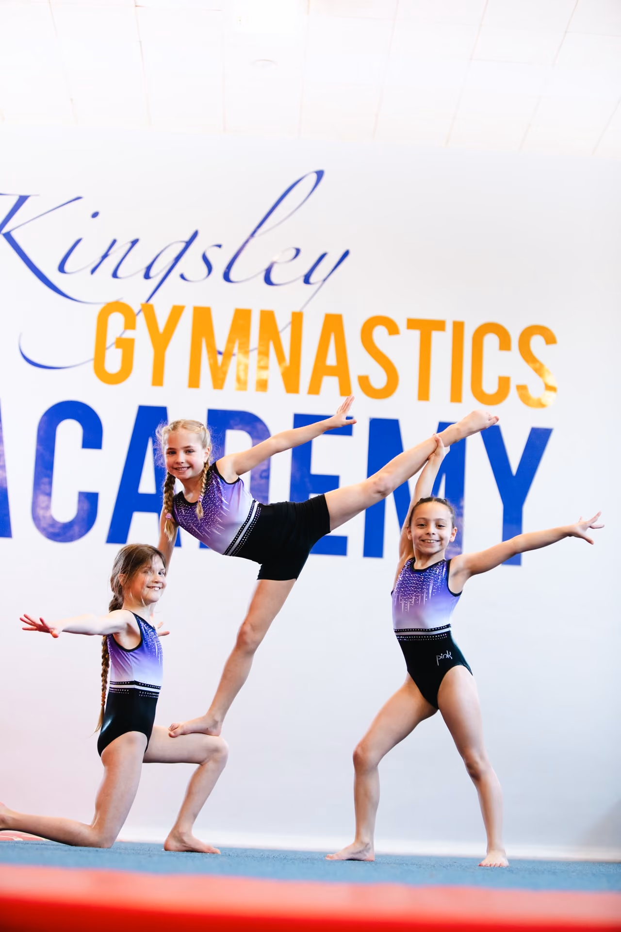 Three young girls in purple and black gymnastics leotards performing poses on a mat in front of a wall displaying the words 'Kingsley Gymnastics Academy'.