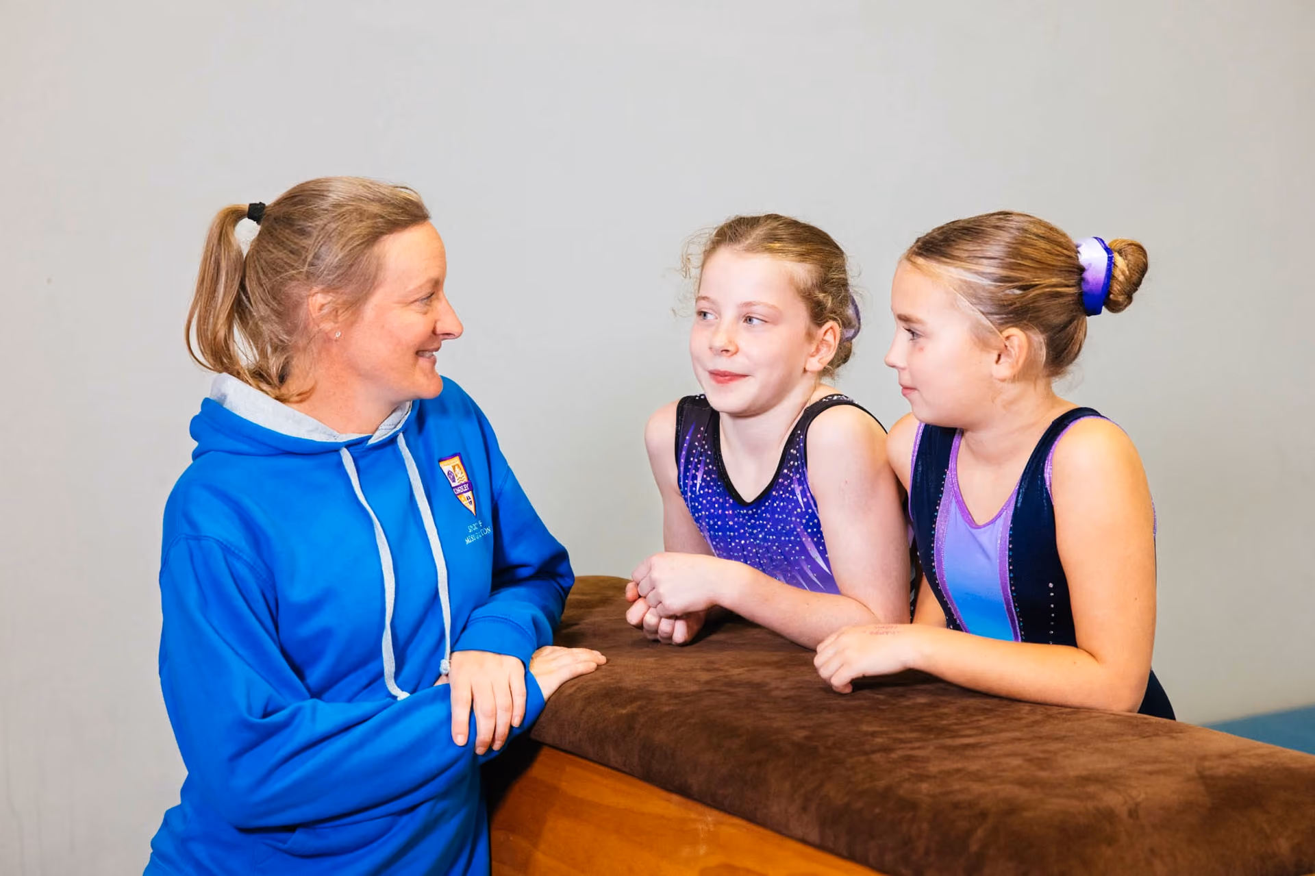 A female coach in a blue hoodie smiles and talks with two young female gymnasts wearing purple and blue leotards, leaning on a padded wooden gymnastics vault.