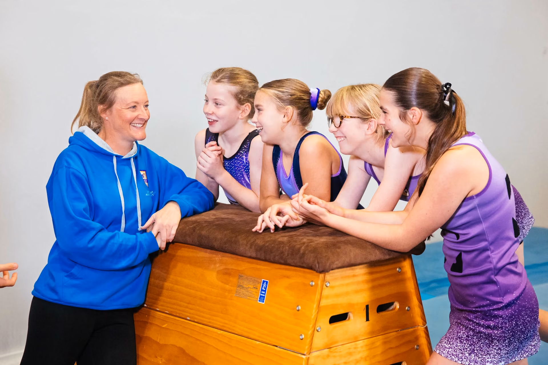 A coach in a blue hoodie talks and smiles with four young female gymnasts in purple leotards leaning on a padded wooden vaulting horse.