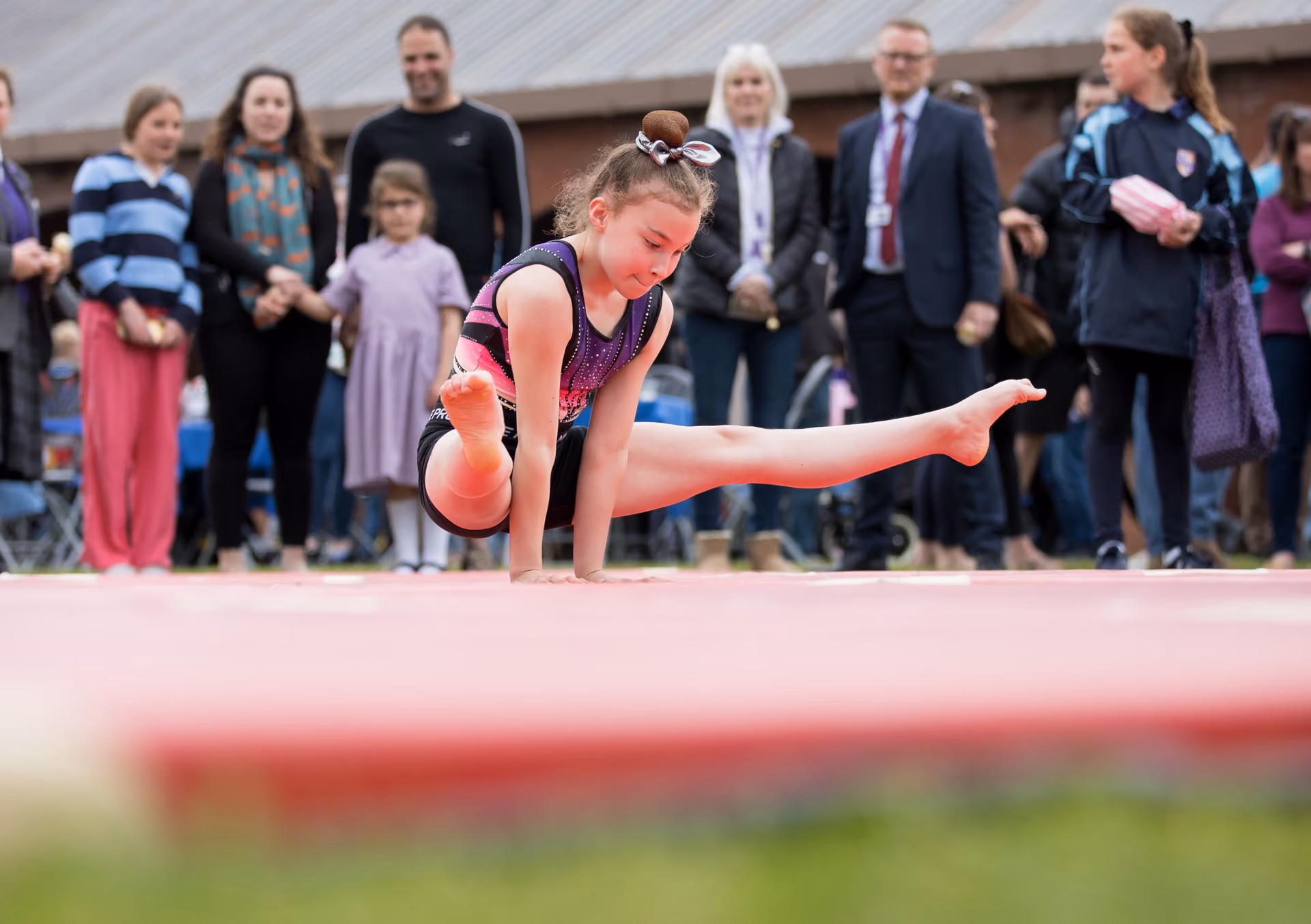 Young girl in a gymnastic leotard performing an arm balance on a pink mat outdoors with a group of people watching in the background.