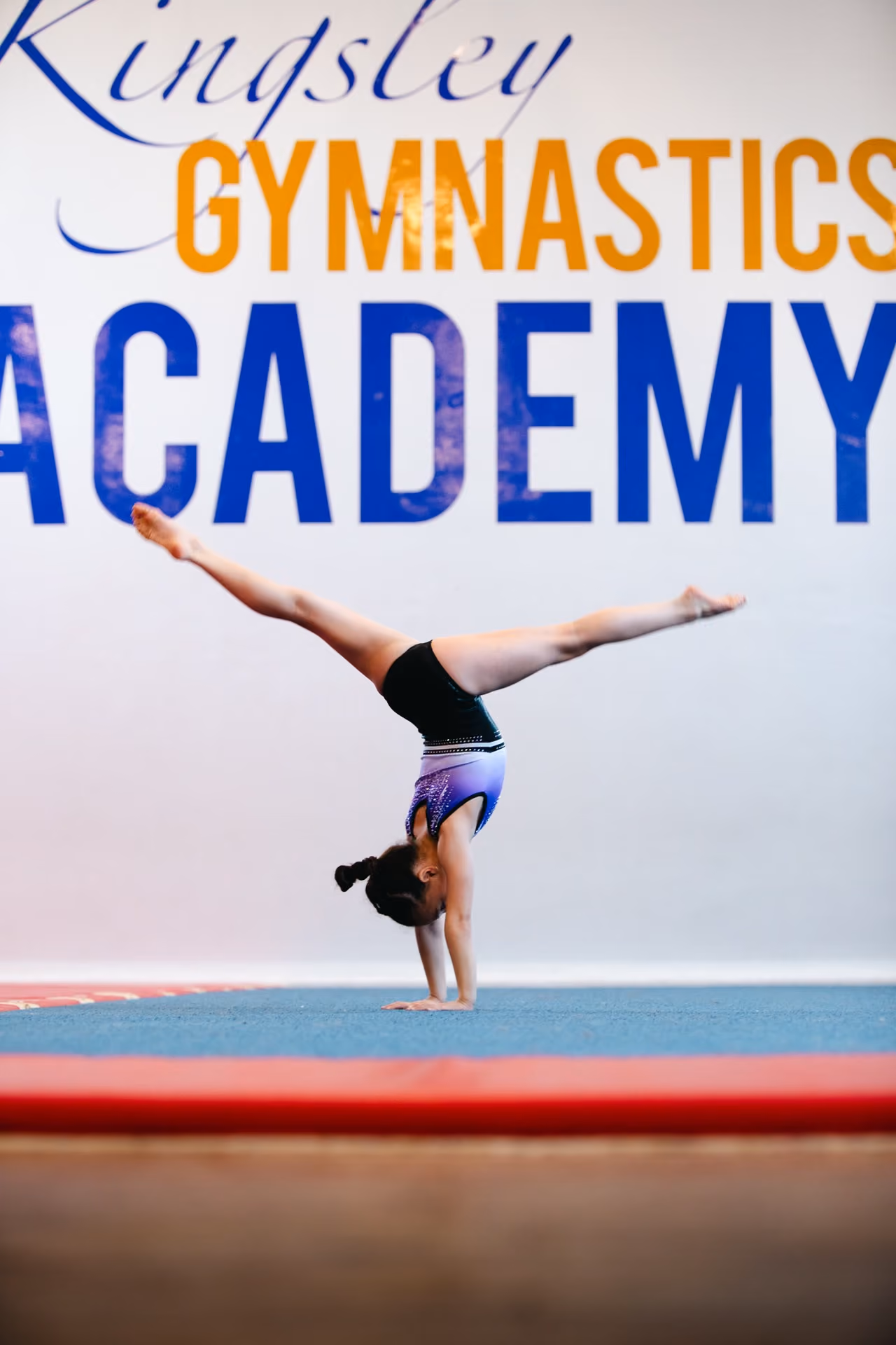 A young gymnast performs a handstand split inside a gymnastics academy with large text on the wall reading 'Kingsley Gymnastics Academy'.