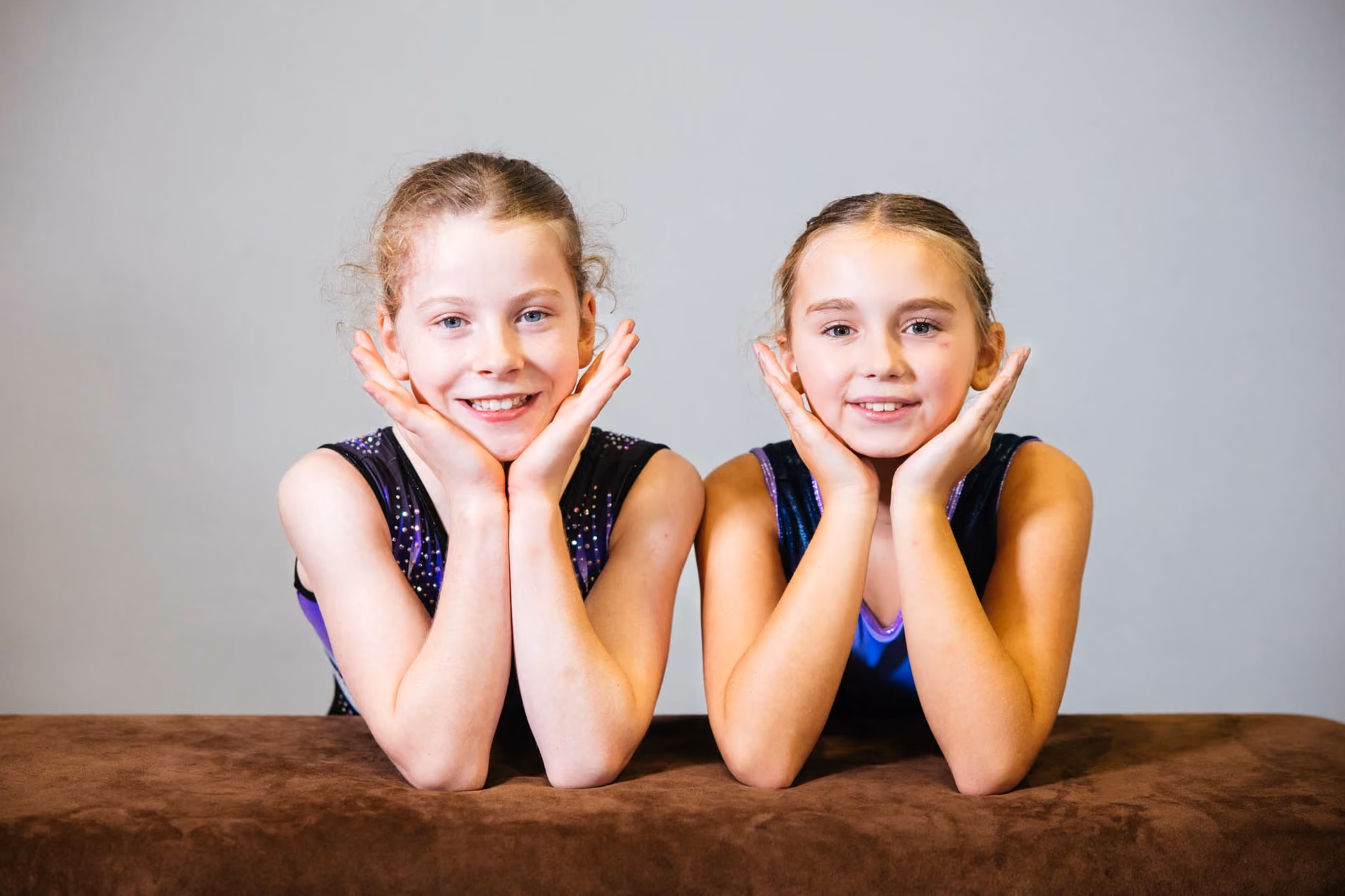 Two young girls in matching sleeveless gymnastics leotards lean on a brown surface, smiling with their chins resting on their hands against a plain grey background.