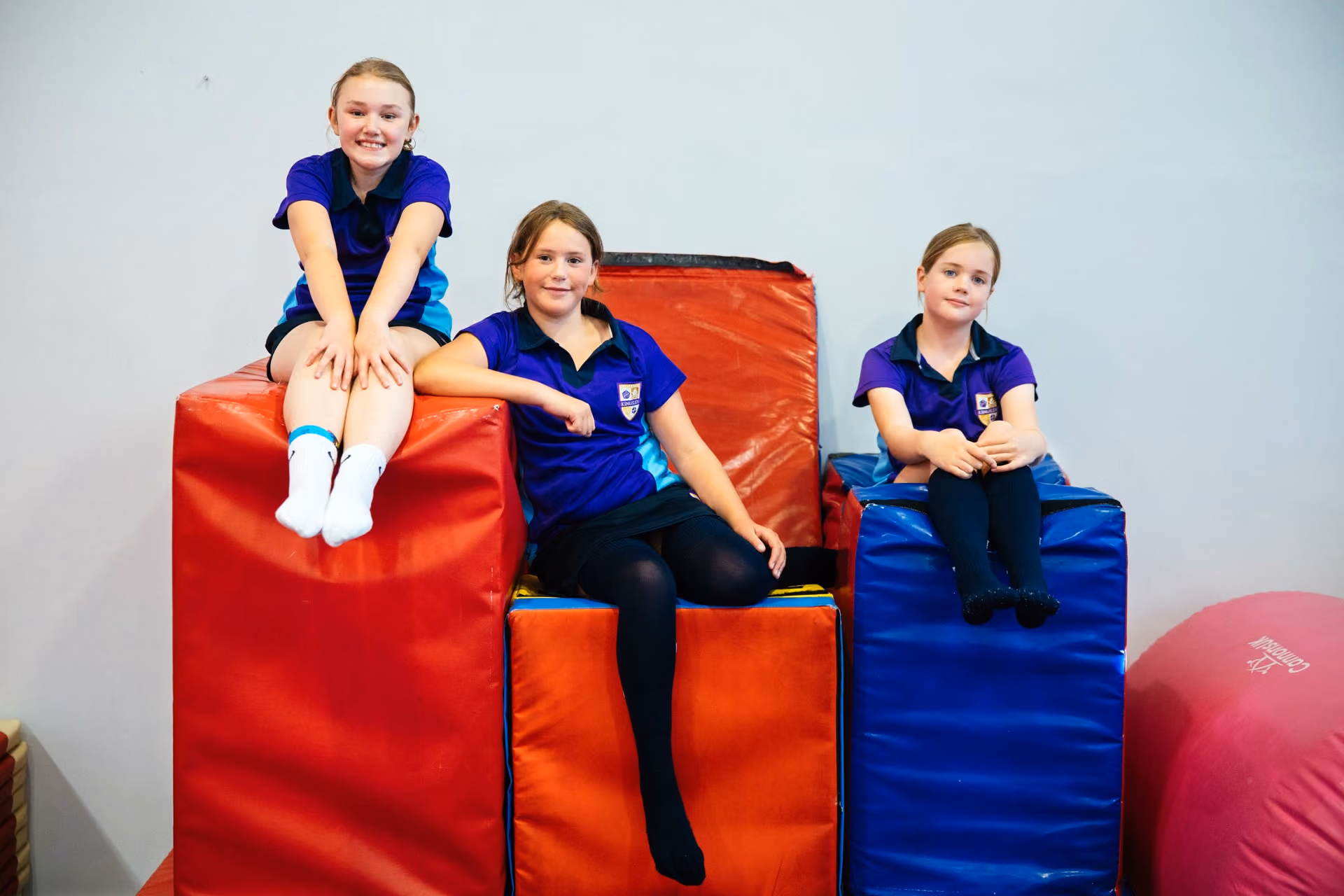Three girls in blue and purple school sports uniforms sitting on large padded blocks in red, orange, and blue inside a gym or activity room.