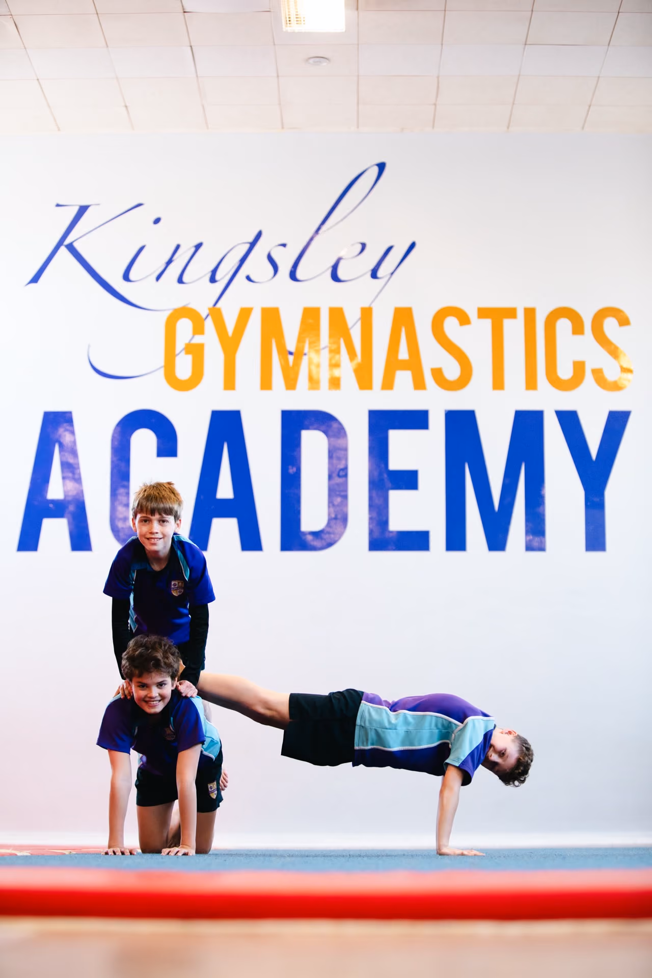 Three children performing a gymnastics pose inside Kingsley Gymnastics Academy with a large wall sign behind them; one child is on hands and knees, another stands behind him, and the third supports himself horizontally with one hand on the floor.