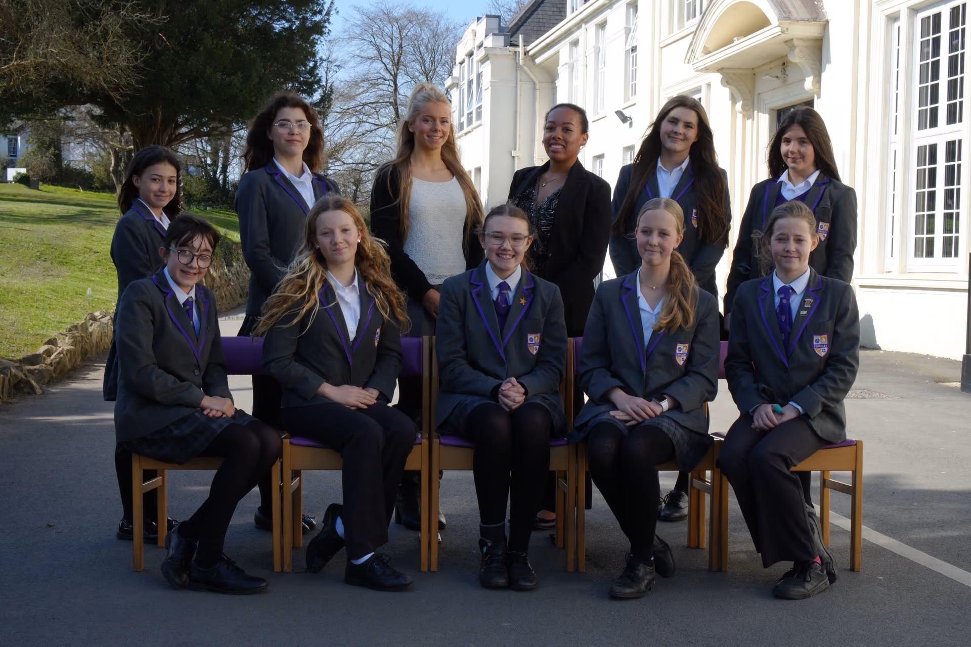 A group of eleven smiling girls and young women posing outdoors in front of a building, most wearing dark school uniforms with purple ties and badges, with some sitting on chairs and others standing behind them.