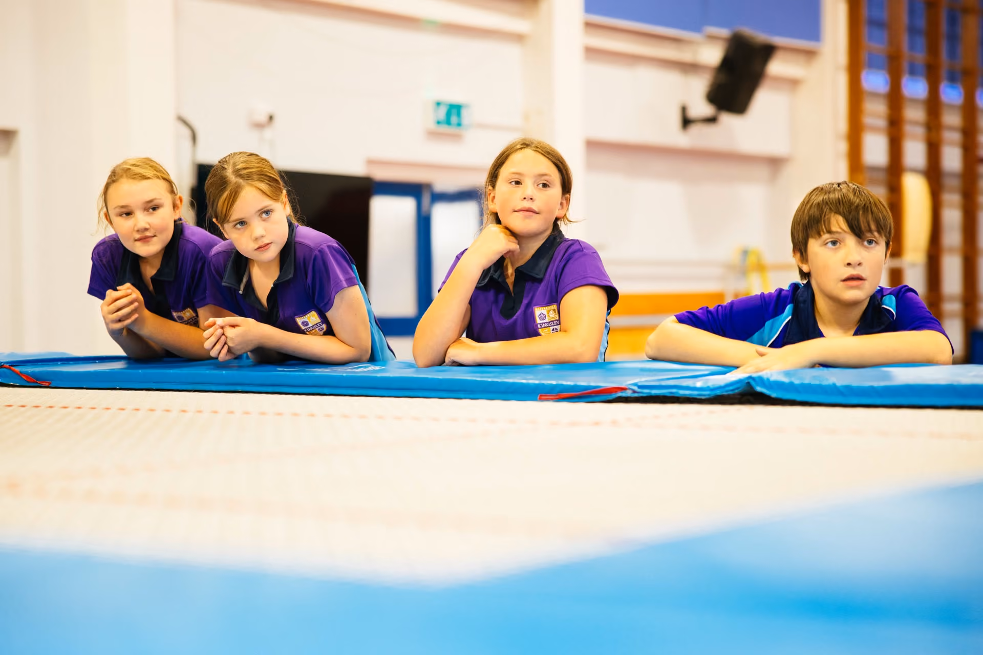 Four children wearing purple sports shirts resting on the edge of a trampoline in an indoor gym, looking attentively in one direction.