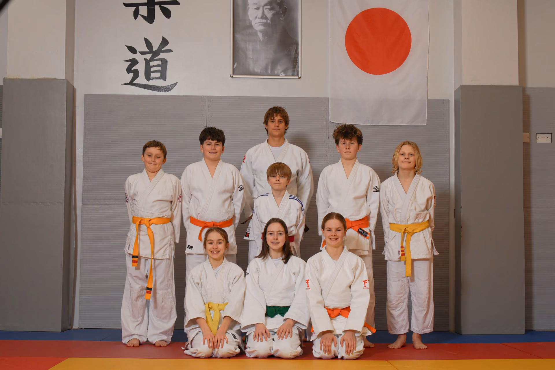 Group photo of nine young judo practitioners posing in white gi uniforms with colored belts, standing and kneeling on mats in a dojo with Japanese calligraphy, a portrait, and a Japanese flag on the wall behind them.