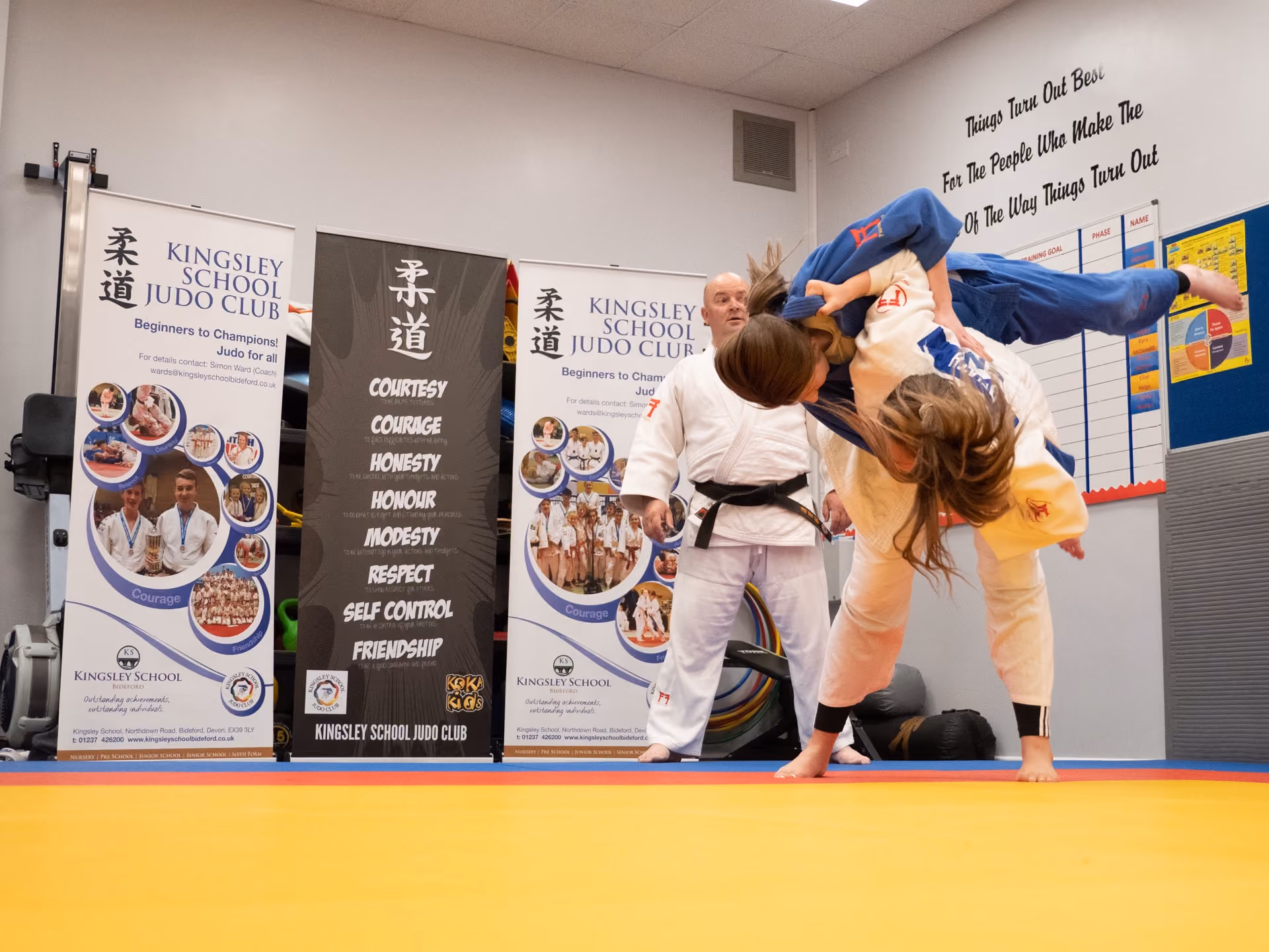 Two female judoka in white and blue gis engaged in a judo throw on a yellow and blue tatami mat, with an instructor in white gi observing in the background inside a Kingsley School Judo Club training room.