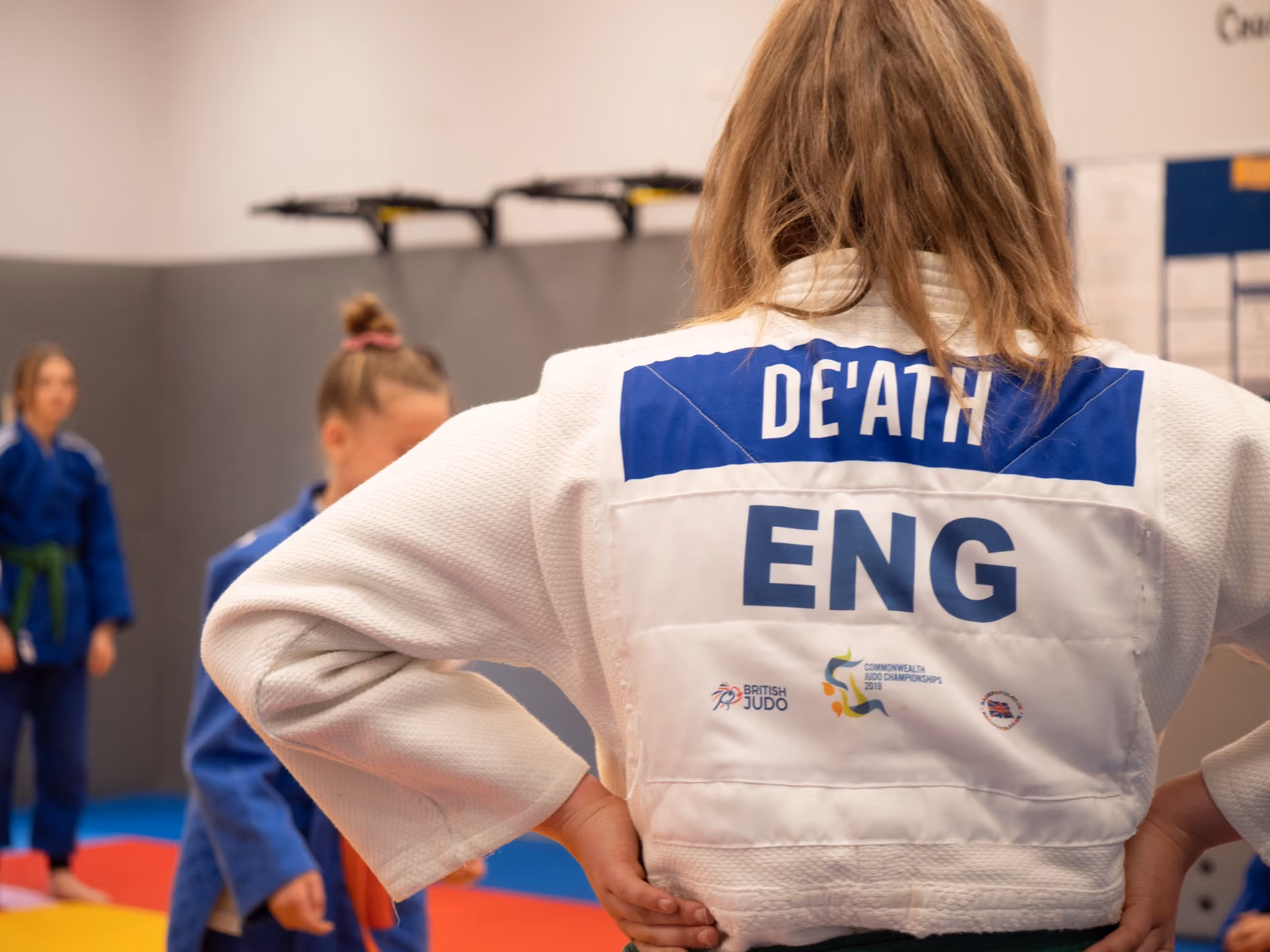 Back view of a judo athlete wearing a white gi with the name 'DE'ATH' and country code 'ENG' on the back, standing on a red and blue mat in a training or competition setting with other athletes in blue gis in the background.