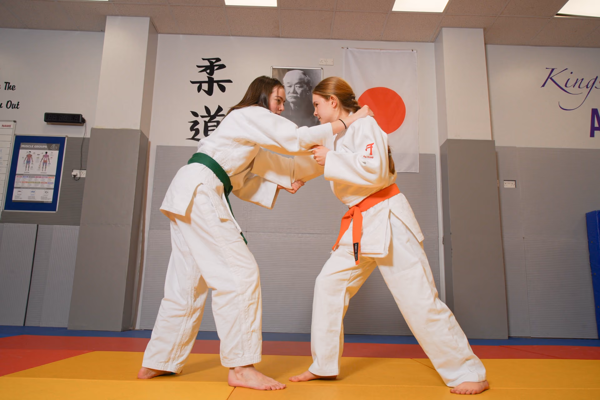 Two women practicing judo in a dojo, wearing traditional white judo uniforms with colored belts, one green and one orange, gripping each other in a grapple stance on a yellow and red mat.