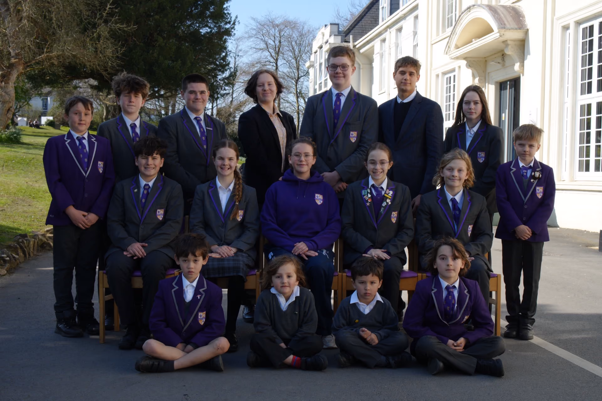 Group photo of a school team with 19 children and one adult standing and sitting outside a white building on a paved area, most wearing purple blazers and school uniforms.