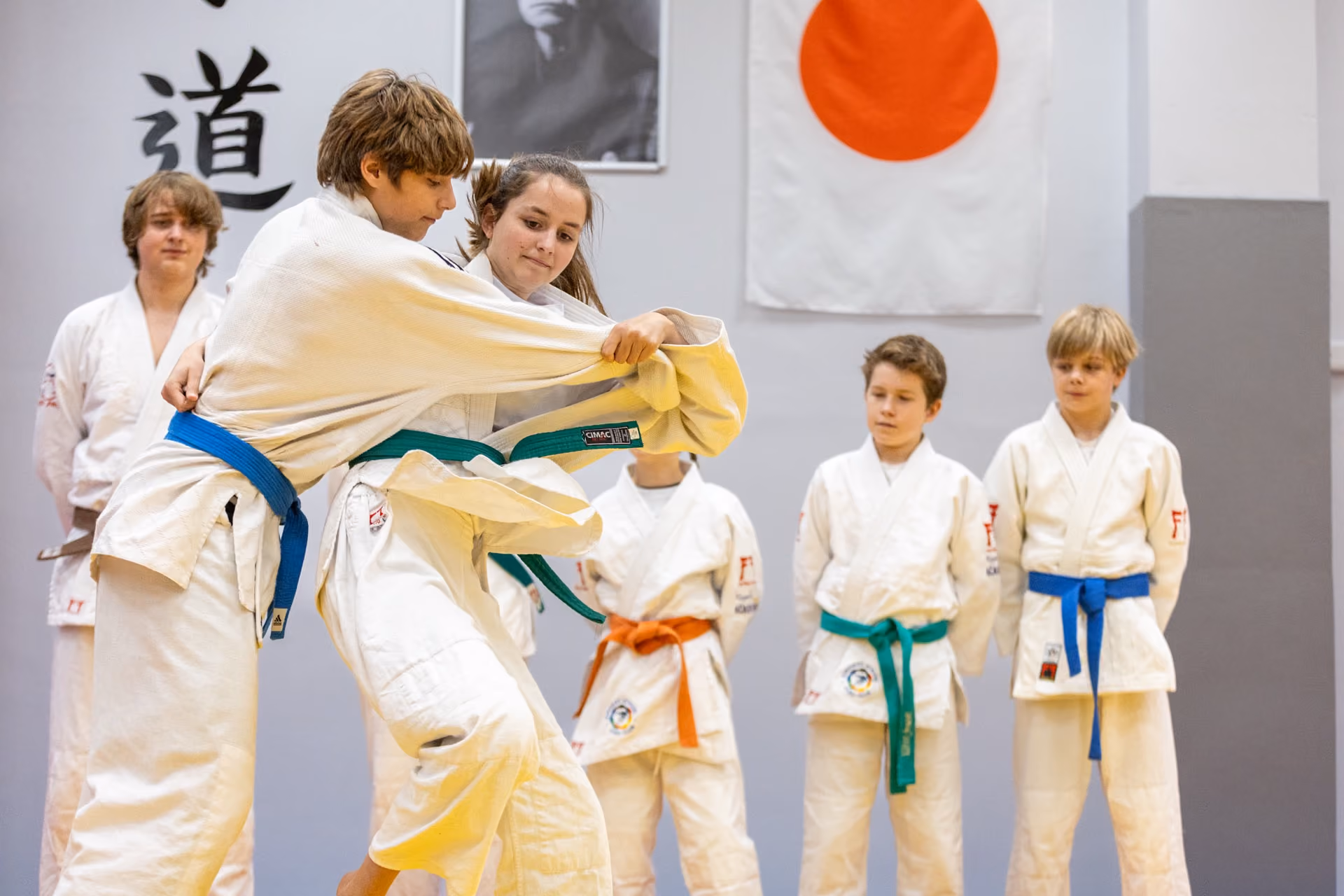 Two young judo practitioners in white gi with blue and green belts engaged in a grappling technique during a training session, while four other children in judo uniforms with various belt colours watch from the background, with a Japanese flag and calligraphy visible on the wall.