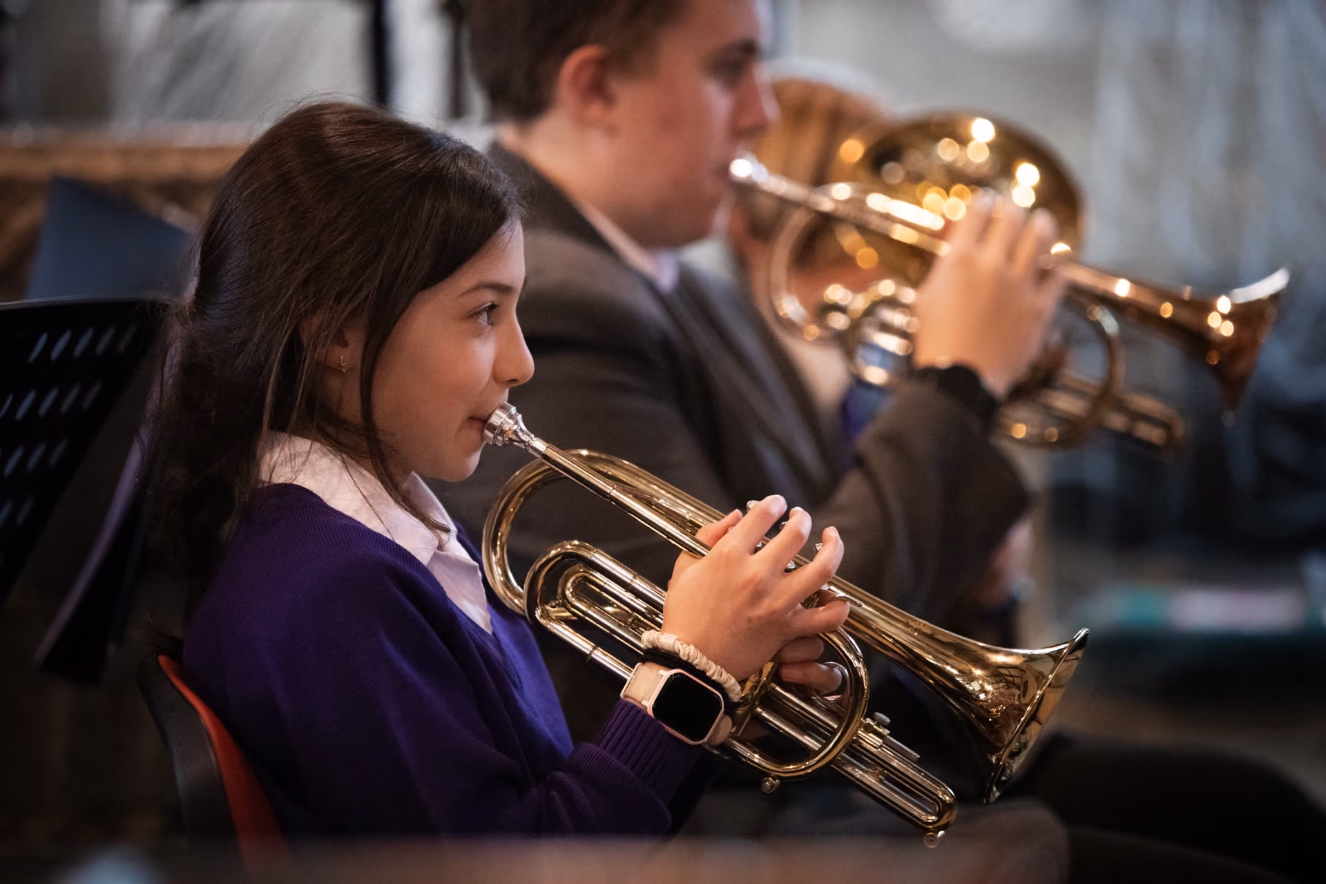 Young girl in a purple sweater playing a brass trumpet, seated next to a boy also playing a trumpet in the background.