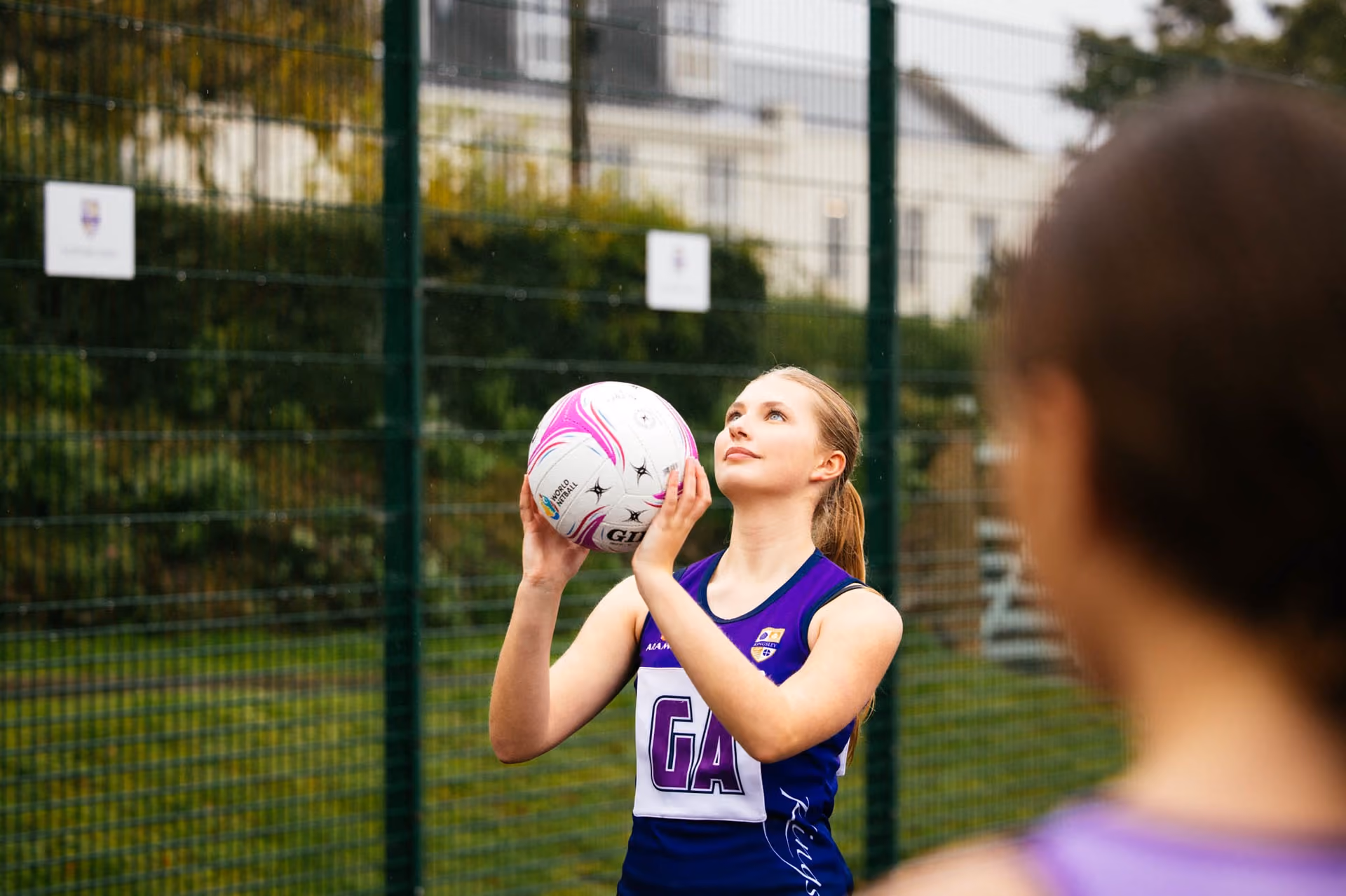 A young woman in a purple netball uniform with 'GA' on the front prepares to shoot a netball on an outdoor court, with another player blurred in the foreground.