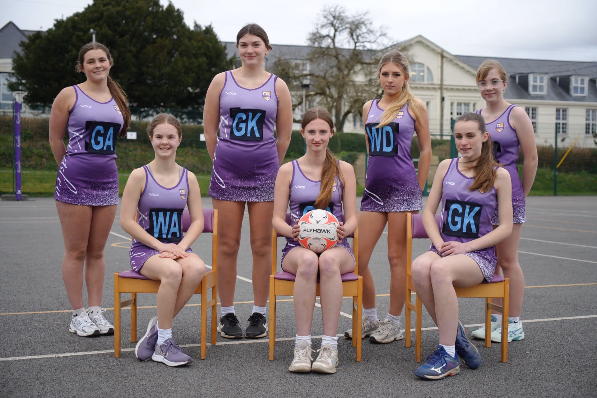 Seven teenage girls in purple and white netball uniforms posing on an outdoor court, three seated on chairs and four standing, with one girl holding a netball with a Flyhawk logo.