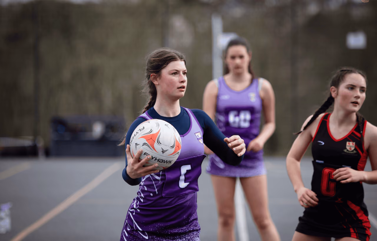 Female netball player in a purple uniform holds the ball with both hands, poised to pass during a game, with two other players in black and purple uniforms in the background on an outdoor court.