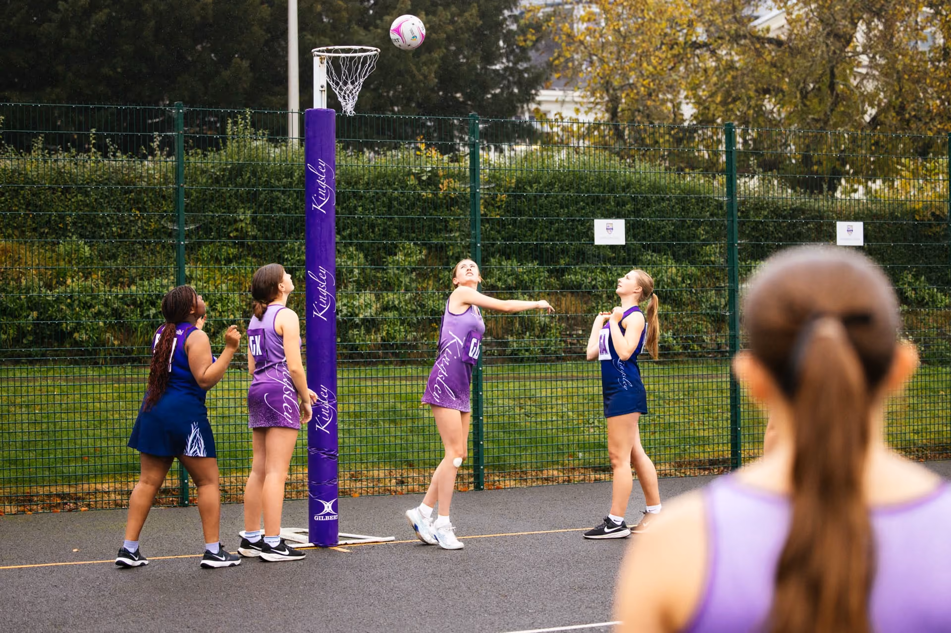 Young female netball players in purple and blue kits playing outdoors on a fenced court, one player in purple is jumping to shoot the ball towards the netball hoop while others watch closely.