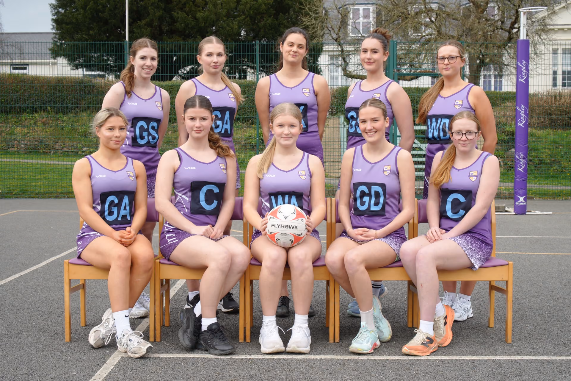 Under-18 netball team posing for a group photo on an outdoor court, wearing purple uniforms with position labels, sitting and standing in two rows, with a netball held by the central seated player.